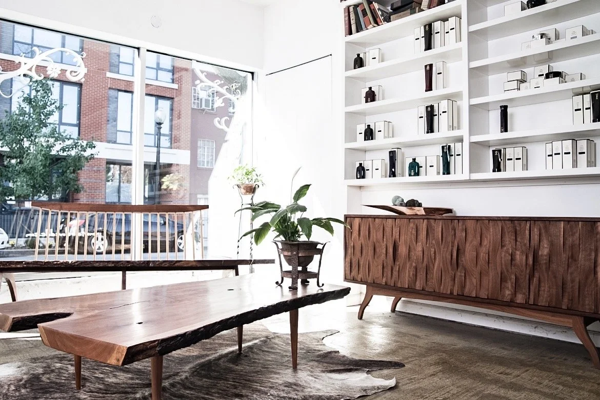 Interior of a modern room with large windows, a wooden coffee table, a sideboard with a textured wood pattern, plants, and white shelves filled with books and decorative objects.