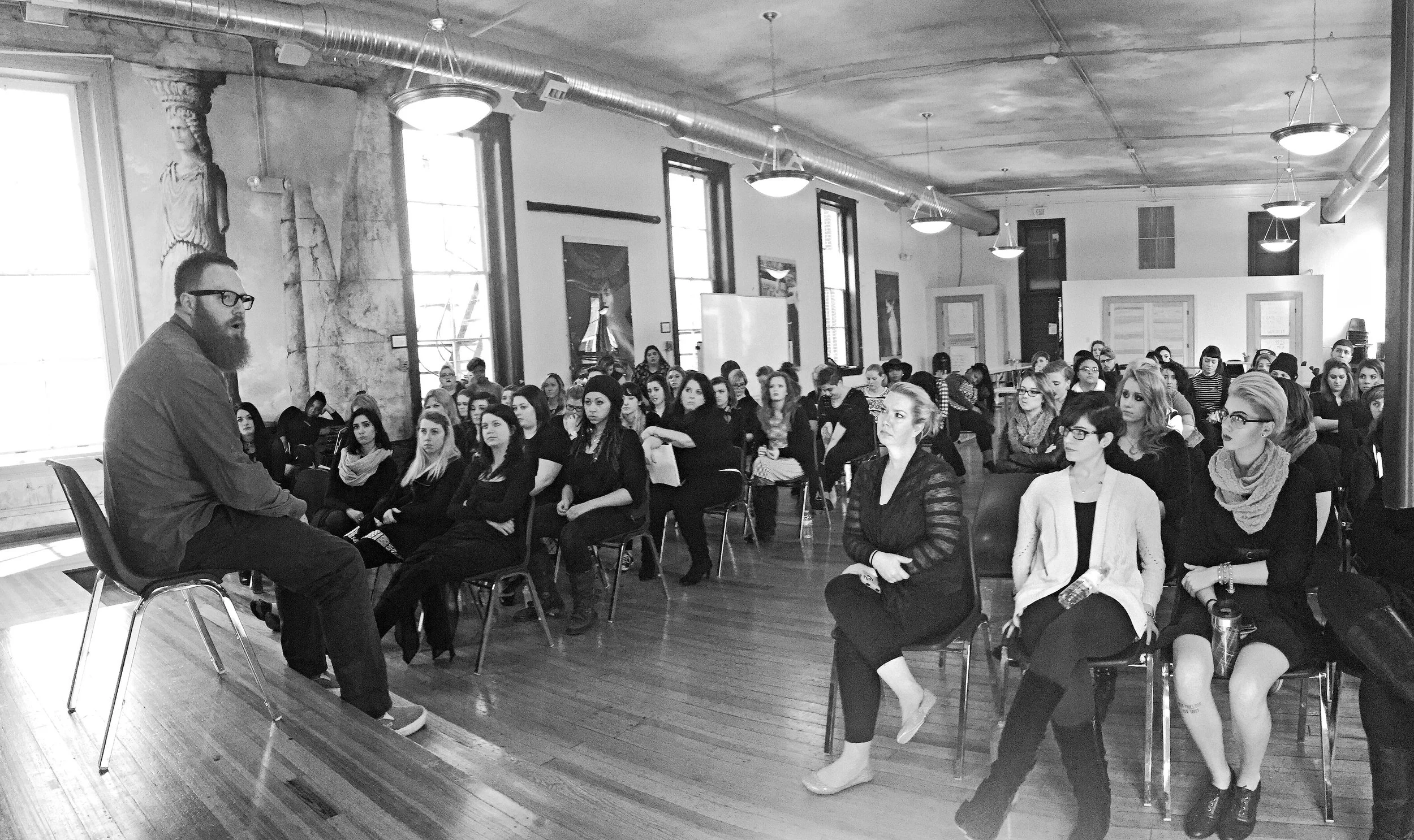 A man with glasses and a beard sits on a chair in front of a group of seated women in a large room with high ceiling, large windows, and artwork on the walls.