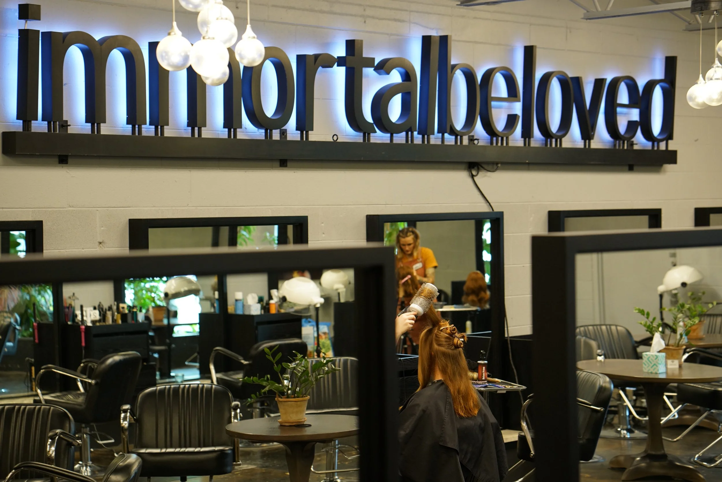 Inside a hair salon with a salon worker styling a customer's hair, sign on the wall reading 'importantbelo', and salon chairs, mirrors, and plants visible.