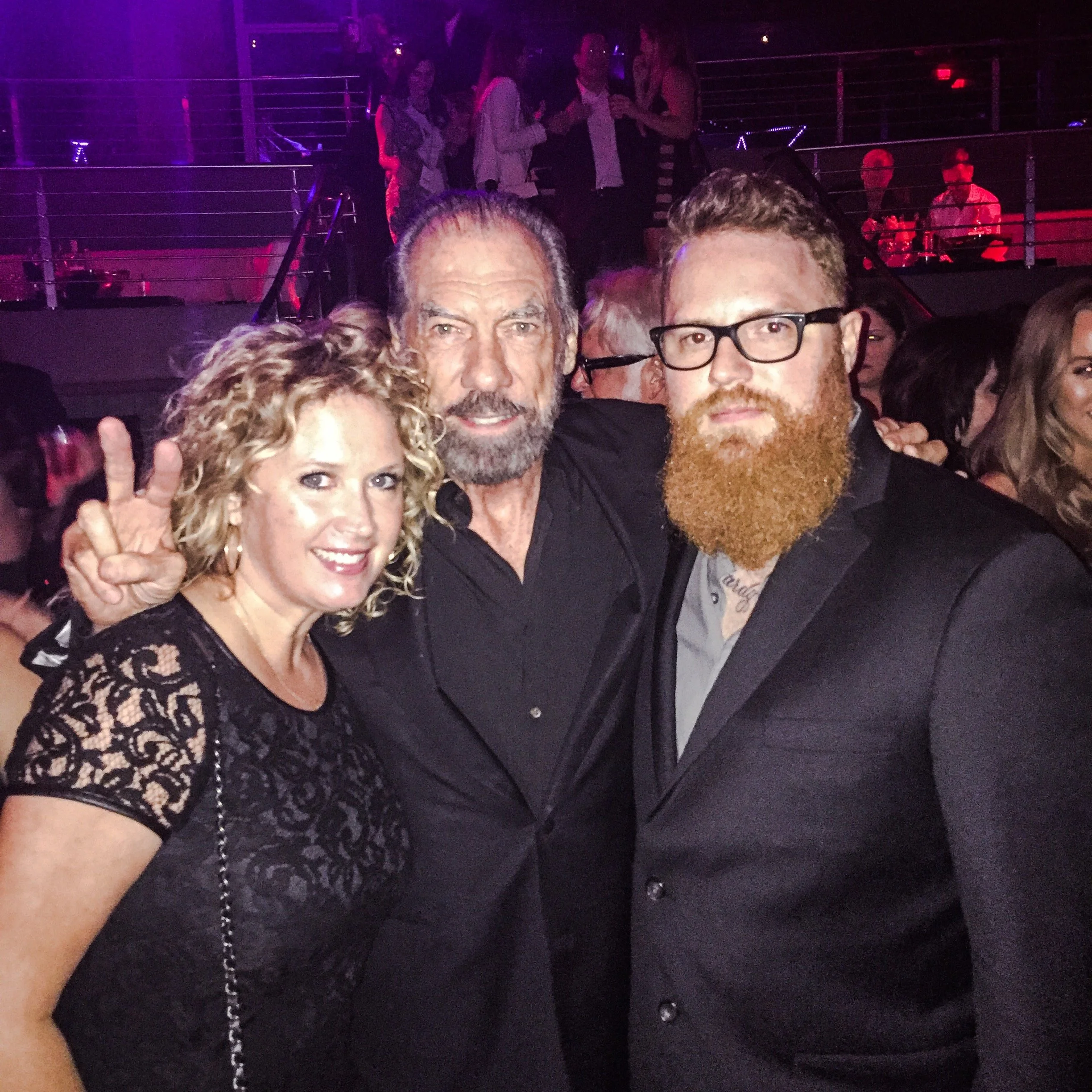 Three people at a party or event, posing for a photo. A woman with curly blonde hair in a black lace dress is on the left, making a peace sign. A bearded man with glasses and a black shirt is in the middle, and a man with a full red beard, glasses, a