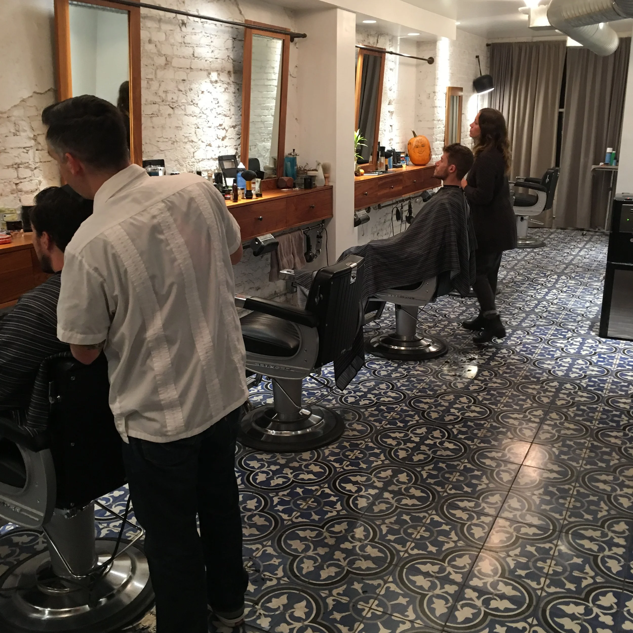 People getting haircuts at a salon with patterned tile floor and white brick walls.
