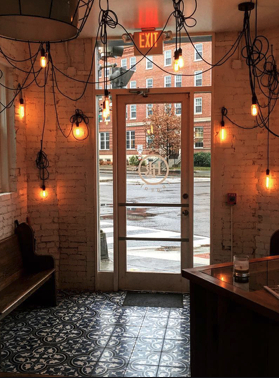 Interior view of a cafe with hanging Edison bulbs, white brick walls, patterned floor tiles, a door with a glass window, and a glimpse of the street outside.