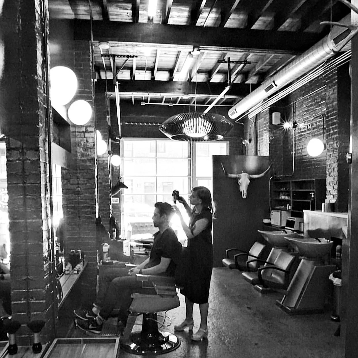A barbershop with a customer sitting in a chair and a barber cutting hair. The shop has exposed brick walls, industrial lighting, and decorative cow skull on the wall.