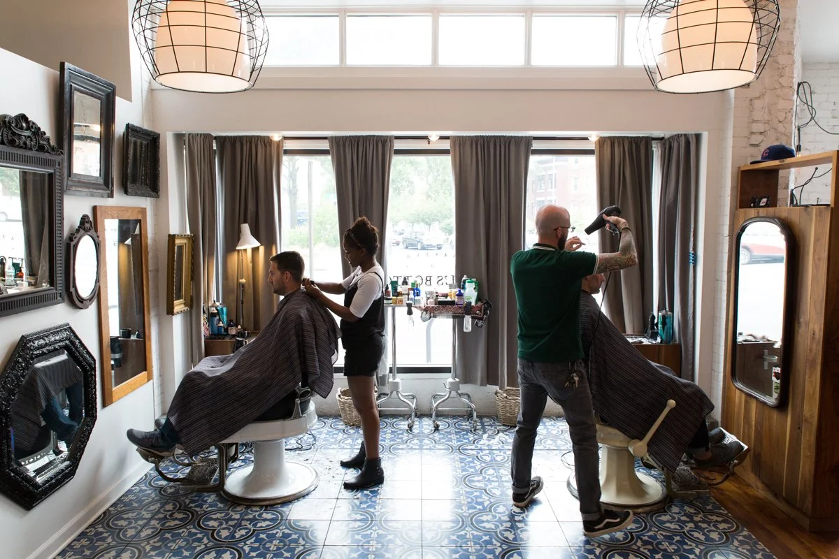 Interior of a vintage-style barbershop with two barbers giving haircuts to clients, mirrors on the wall, framed pictures, and decorative lighting.