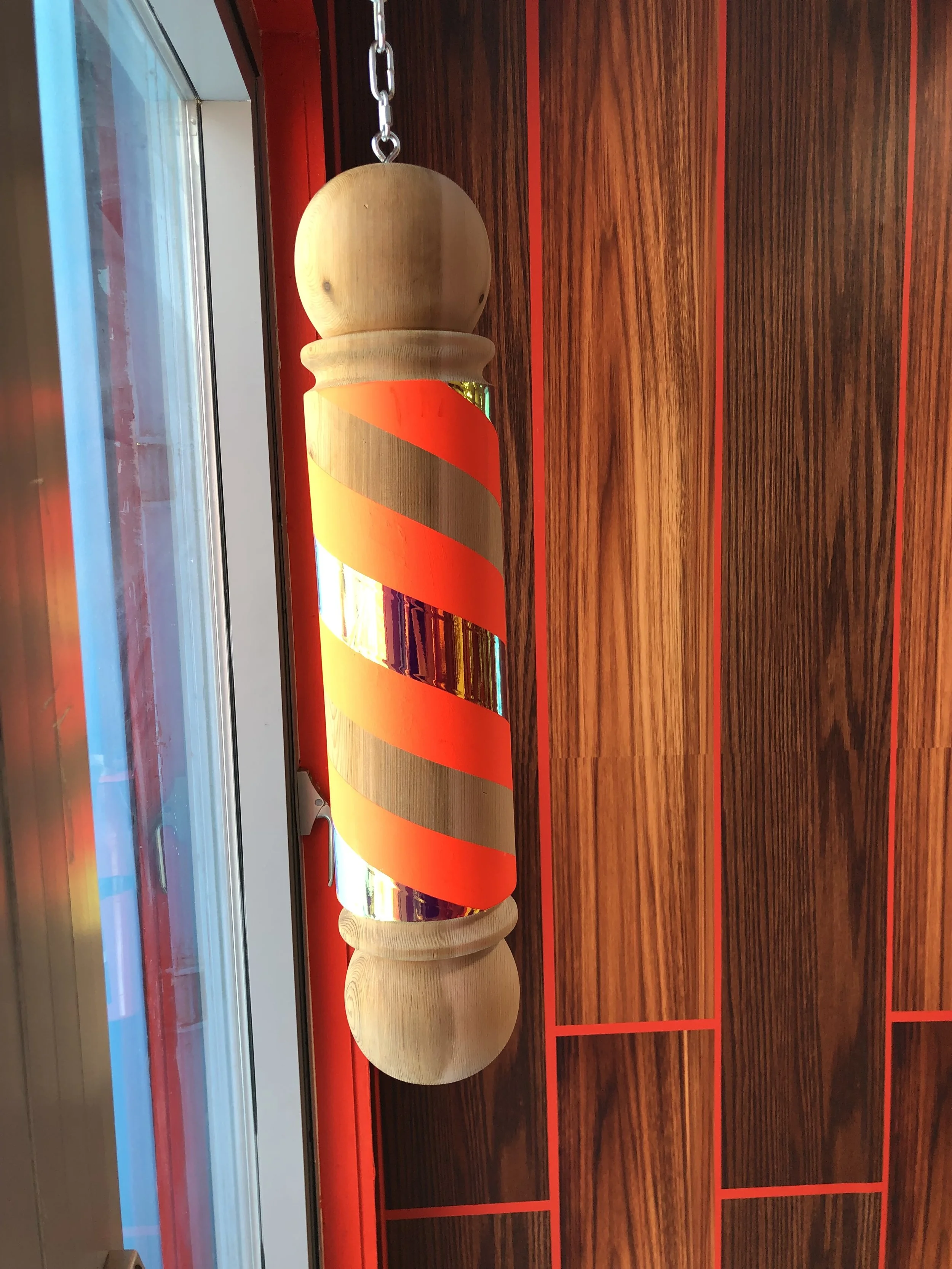 A barber pole with orange and reflective silver stripes on a wooden wall near a window.