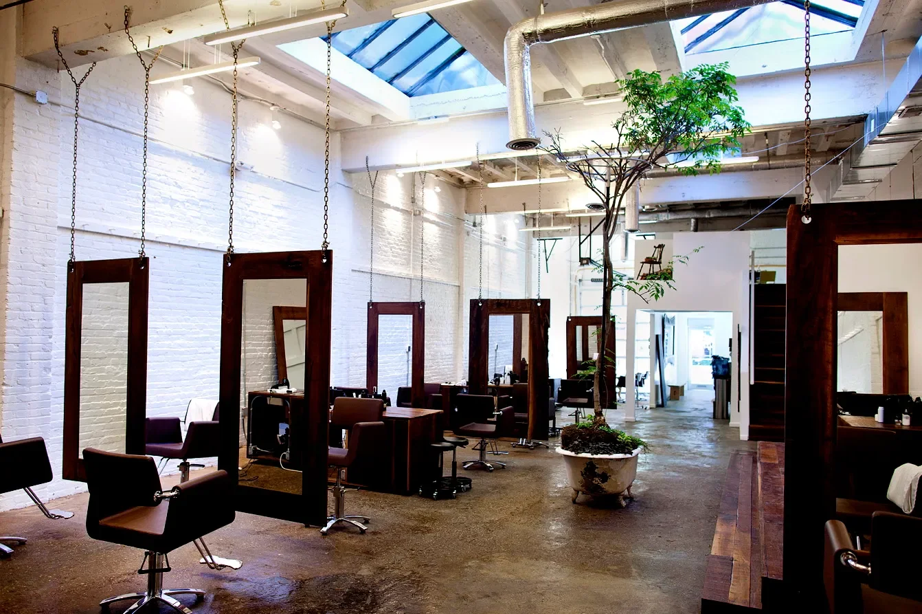 Interior of a hair salon with styling stations, chairs, rectangular mirrors, and a large potted plant in the center; white brick walls and an industrial ceiling with skylights.