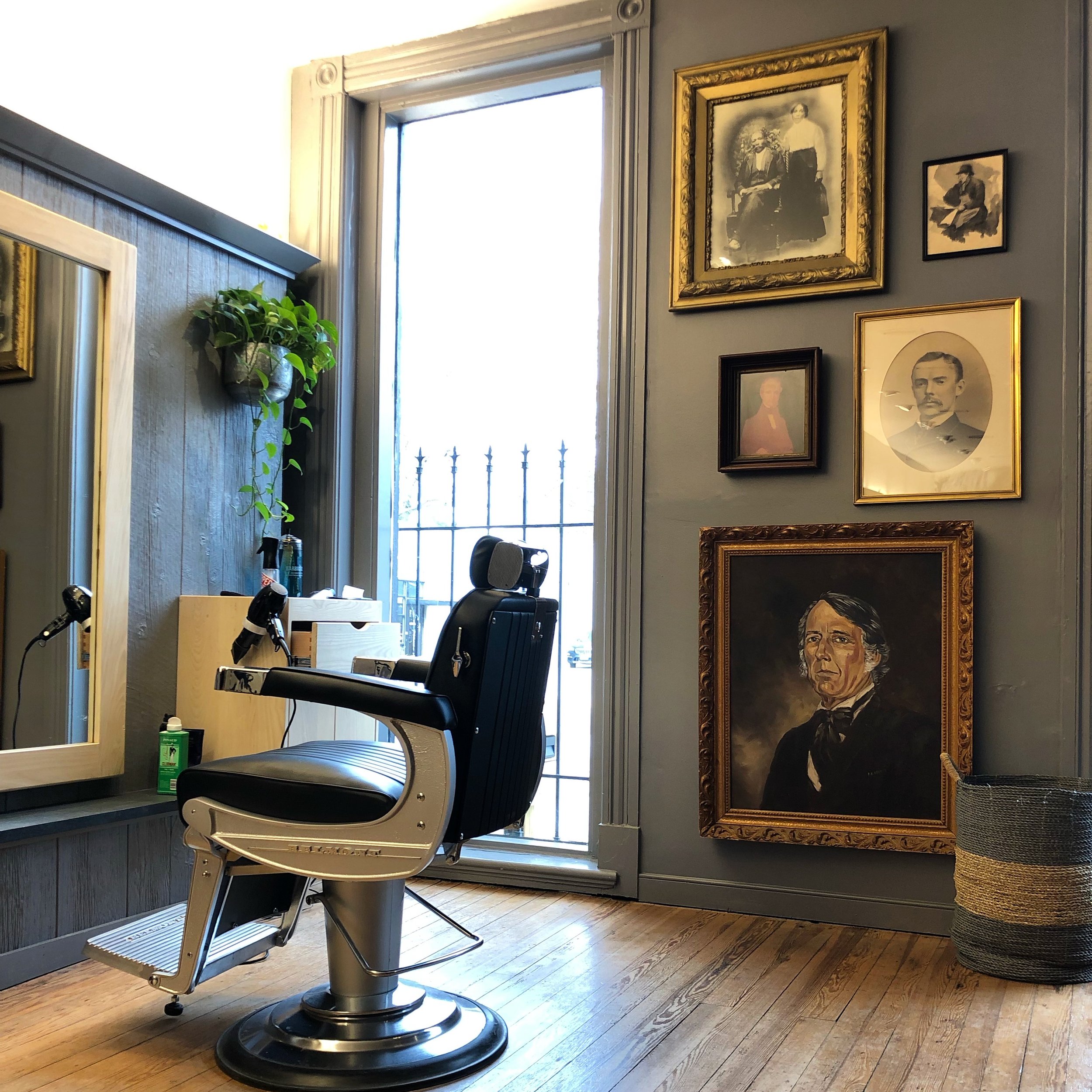 A vintage barbershop with a barber chair, a mirror, and framed portraits and paintings on the wall, illuminated by natural light from a large window.