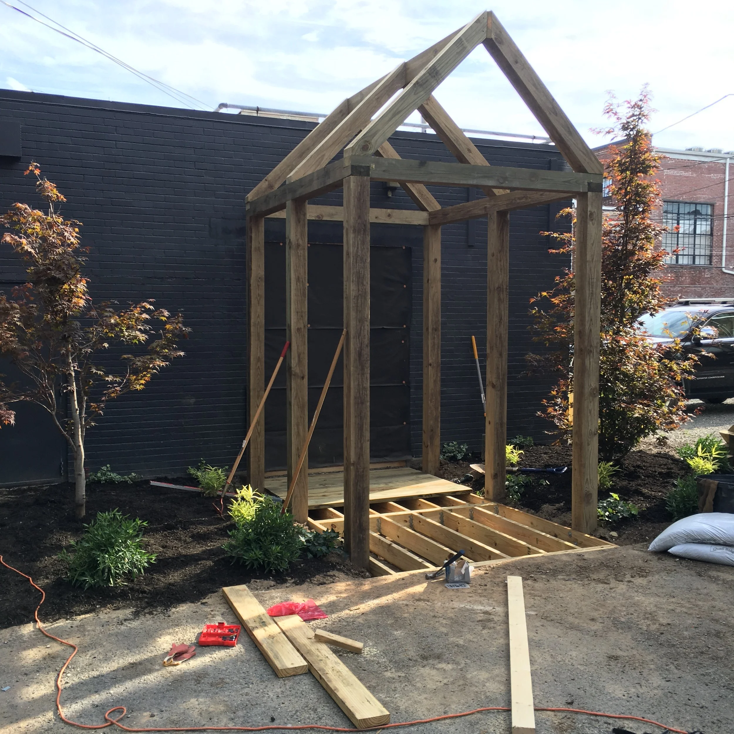 Wooden structure under construction with a garden and trees in the background.