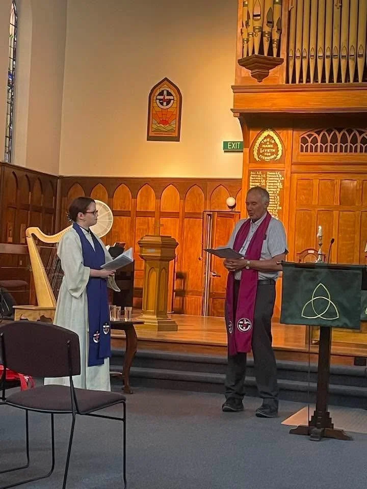 A woman and a man are standing inside a church, holding papers. The woman is dressed in a white robe with a blue stole, and the man is wearing a gray shirt with a pink stole. They are on a stage with wooden paneling and a pipe organ in the background