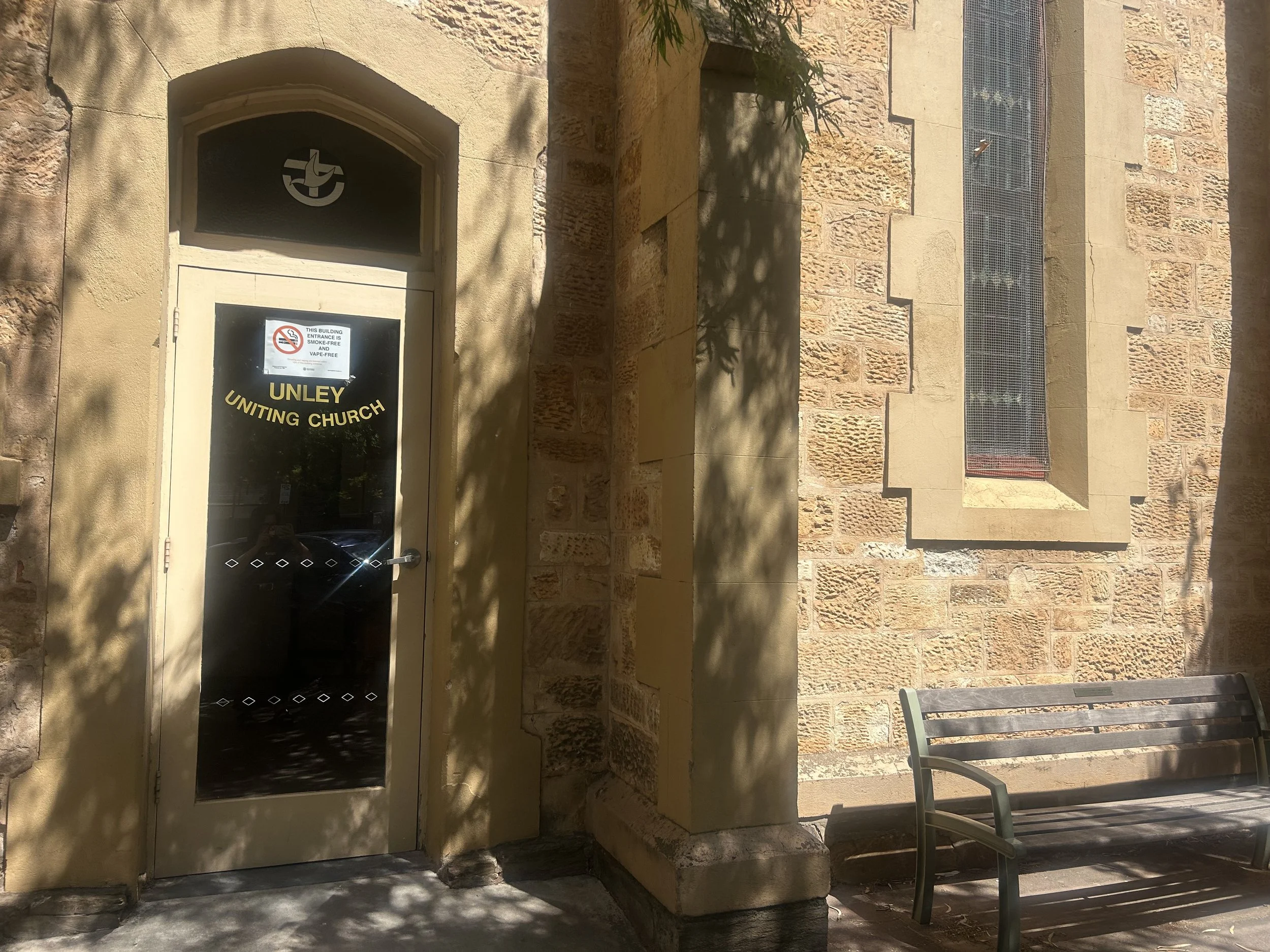 The entrance door of Unley Uniting Church with a sign indicating no smoking or vaping inside. The building has brick walls, a window with bars, and a nearby bench.
