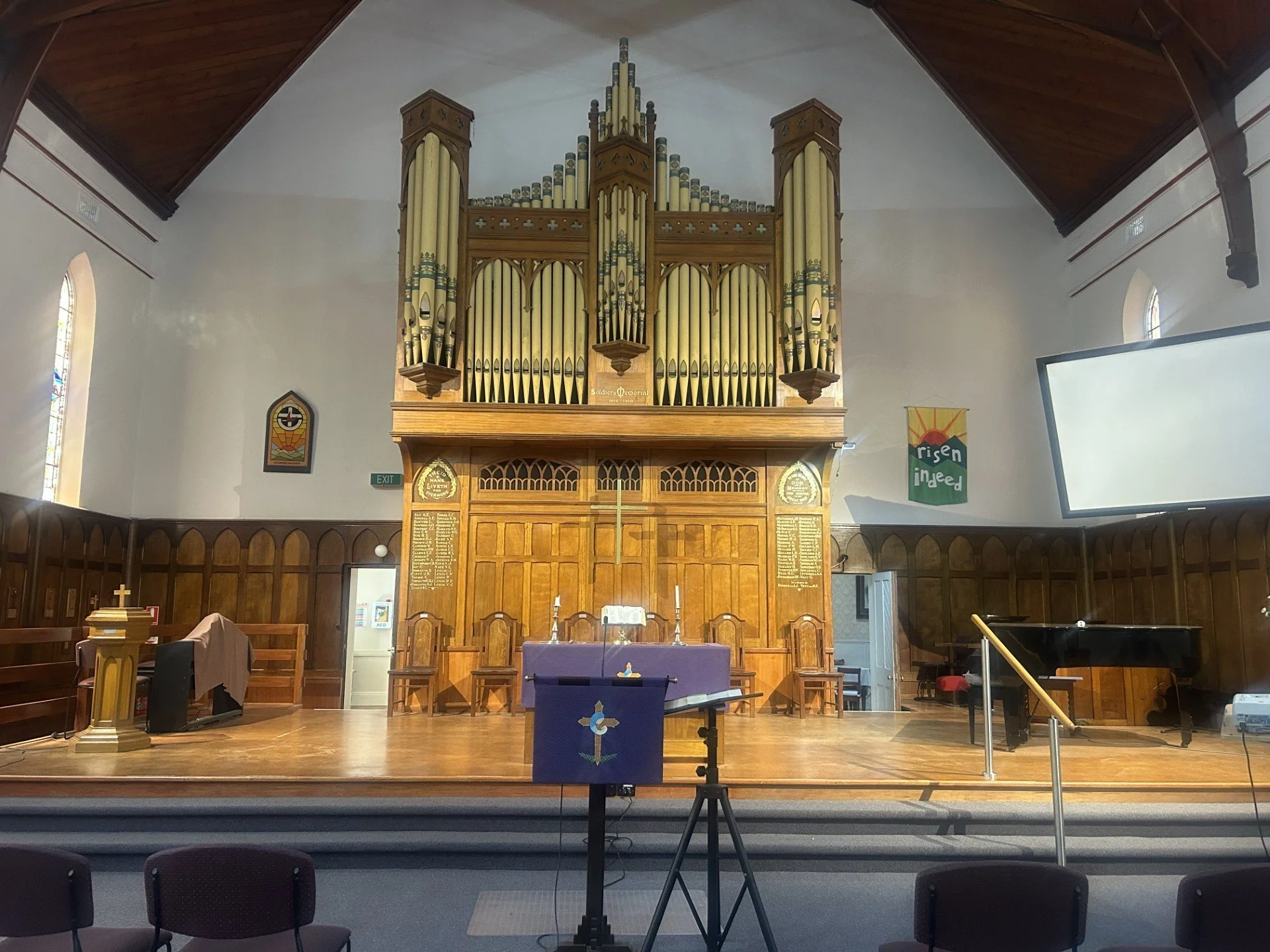 Interior of a church sanctuary with a large pipe organ at the front, wooden pews, a purple altar cloth, a piano on the right, and stained glass windows on the sides.