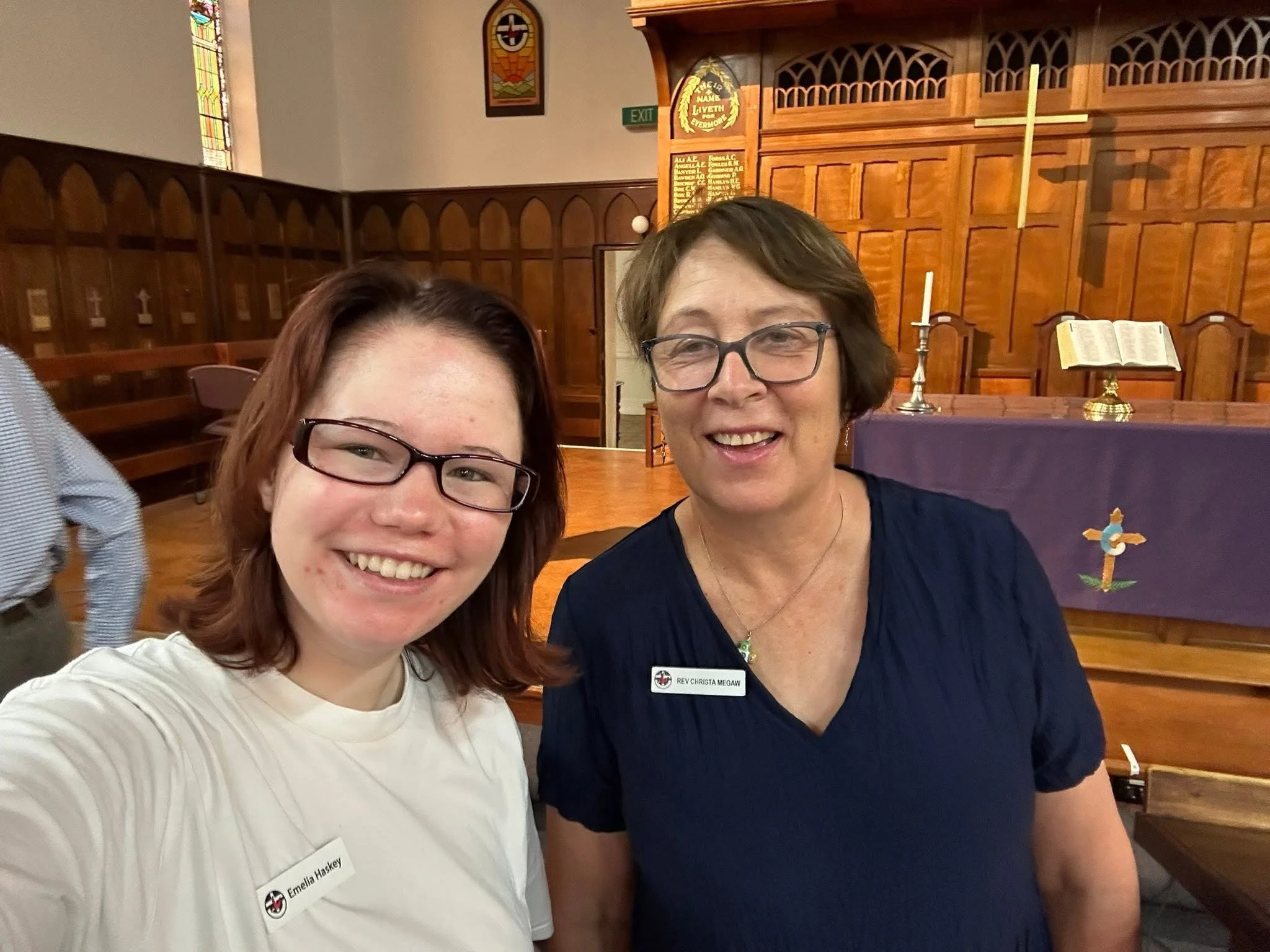 Two women smiling for a selfie inside a church, with wooden walls and religious symbols in the background. One woman is wearing a white shirt and glasses, and the other is wearing a navy dress and glasses.