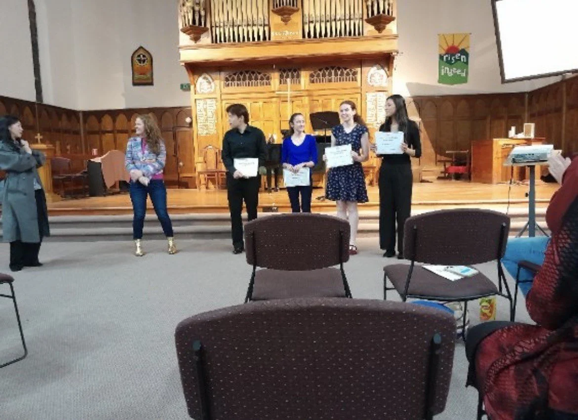 Group of people standing on stage holding certificates in a church or auditorium setting, audience seated in front.