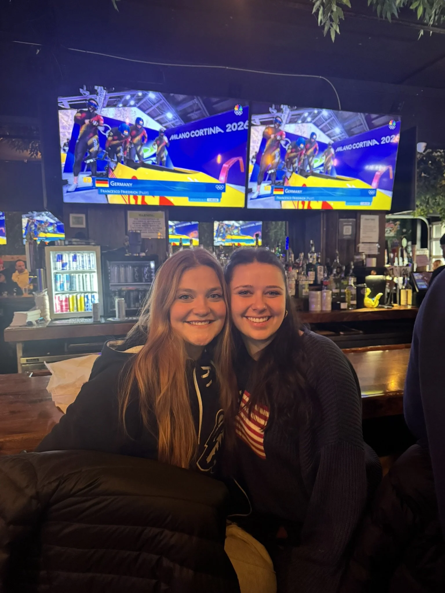 Two women smiling sitting at a bar with multiple TV screens showing a cycling event in Milano Cortina 2026 in the background.