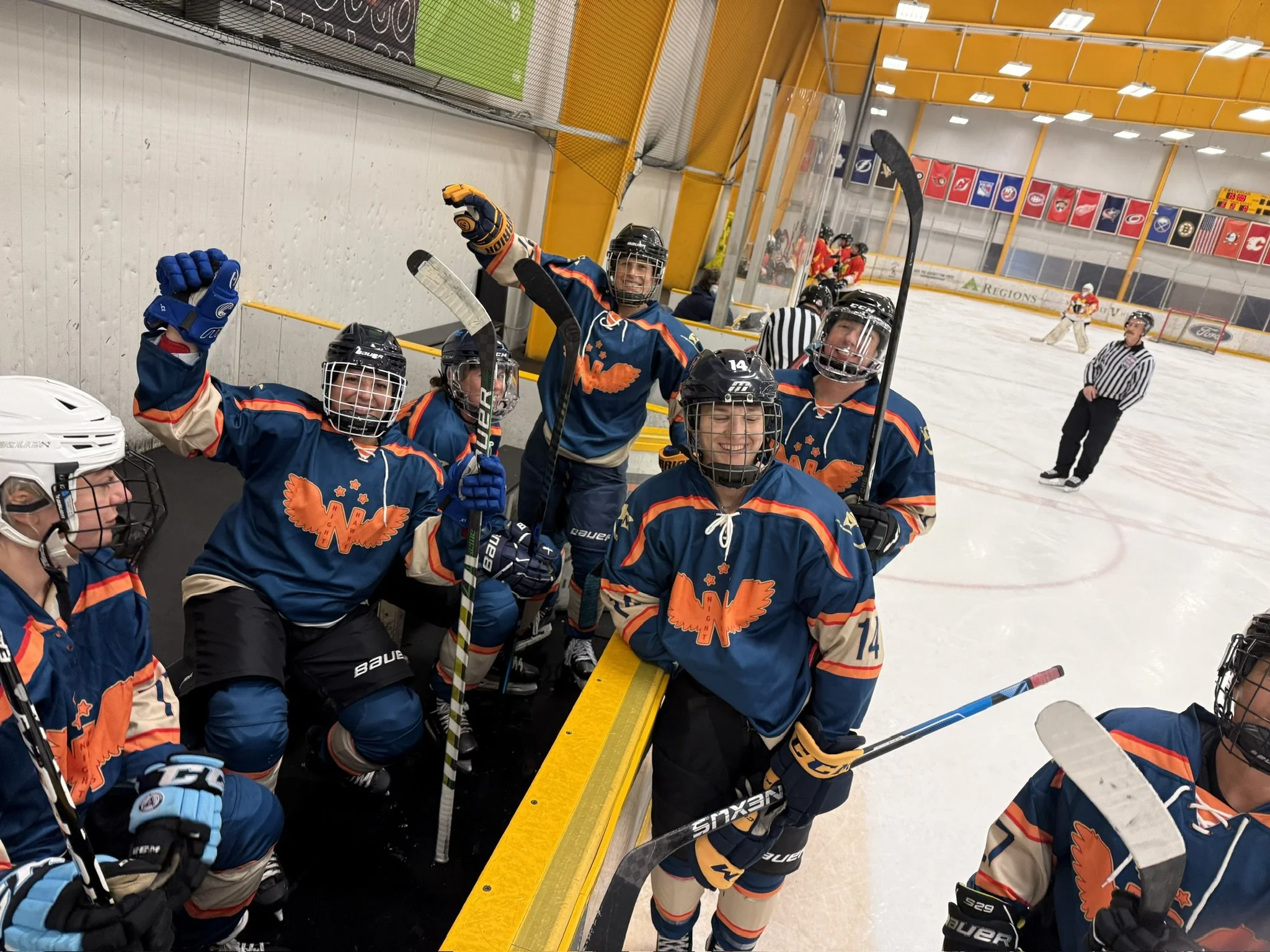 Hockey team in navy blue and orange uniforms celebrating on the bench in an indoor ice rink, with players and referees visible.
