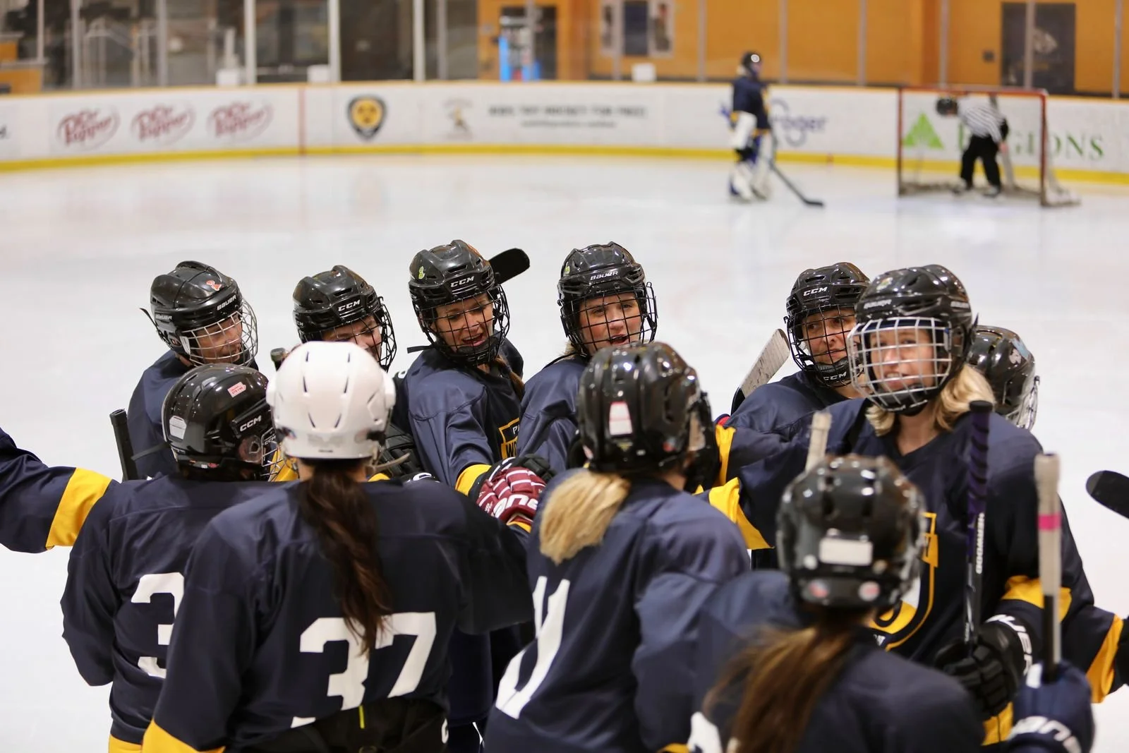 Group of female ice hockey players in dark blue jerseys celebrating on the ice rink, with a referee and a goalie in the background.