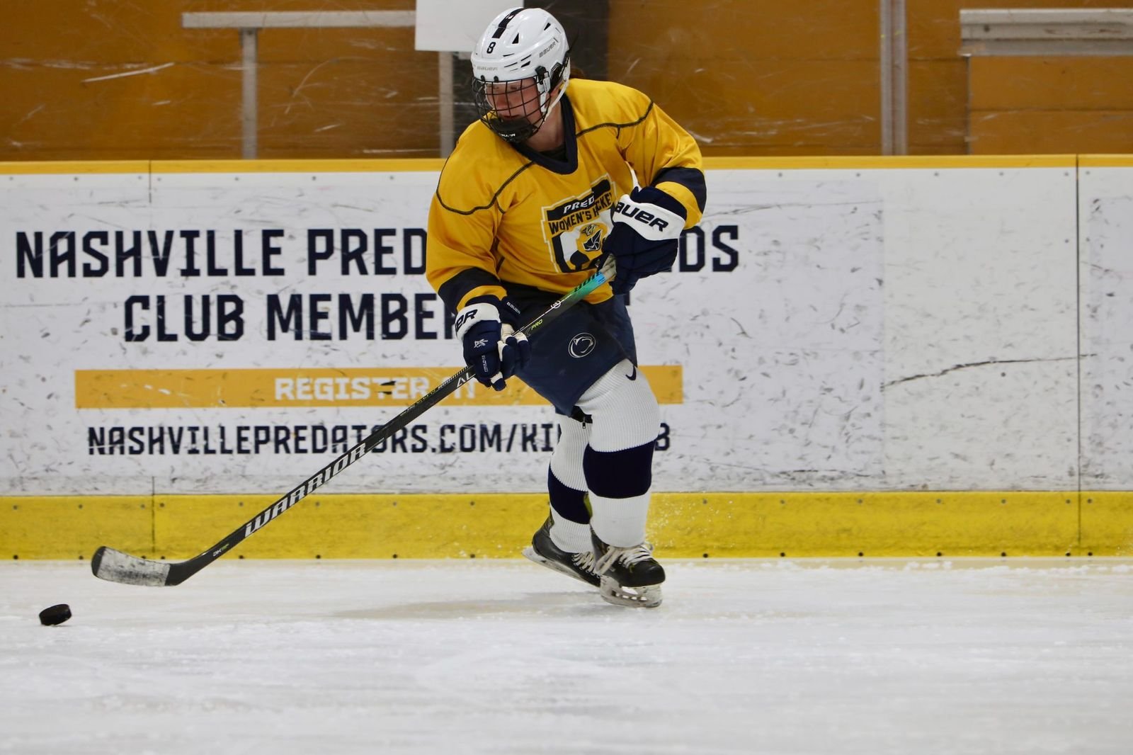 Ice hockey player in yellow jersey, black helmet, and gloves, skating on ice rink with a hockey stick, chasing a puck, with a sign reading "Nashville Predators Club Members" in the background.