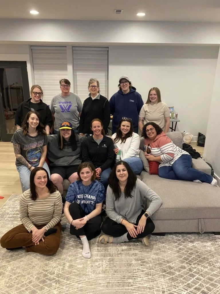 Group of fifteen women gathered in a living room, some seated on the floor, others on a sofa, smiling for a group photo.