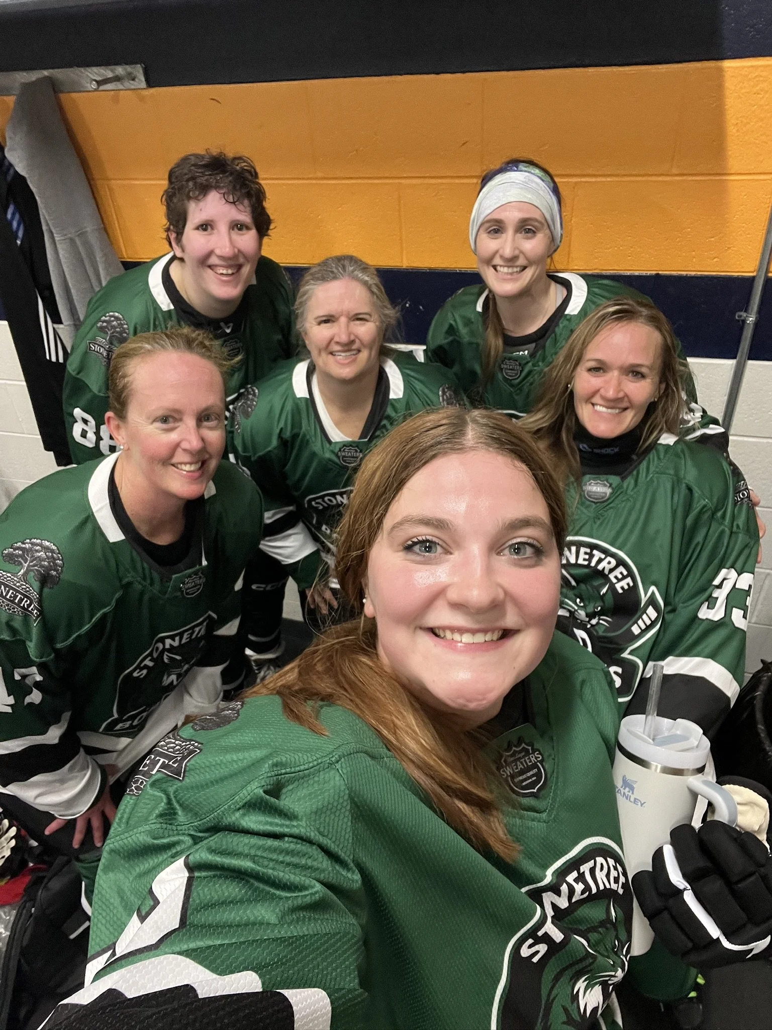 Group of six women hockey players in green jerseys smiling for a selfie in locker room.