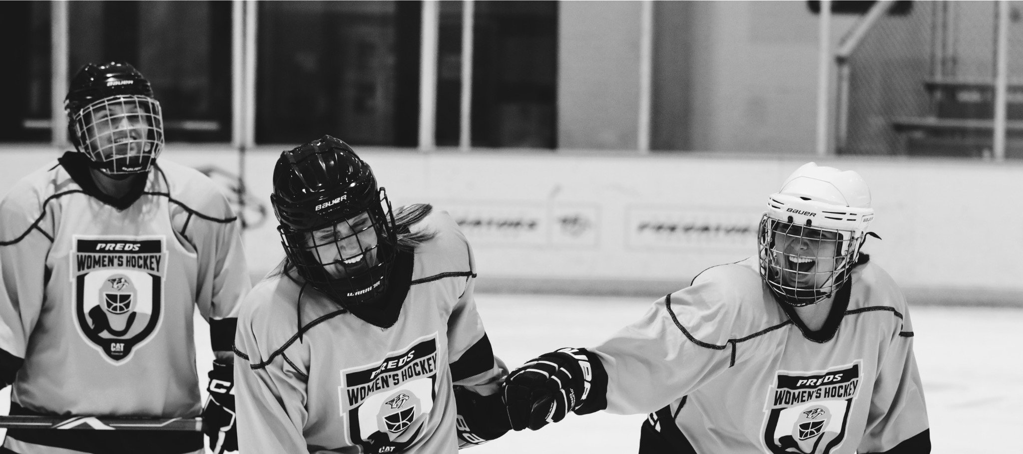 Three women hockey players wearing helmets and matching jerseys with a women's hockey logo on the front, on the ice rink. One player is smiling with their mouth open, two others are in mid-celebration.