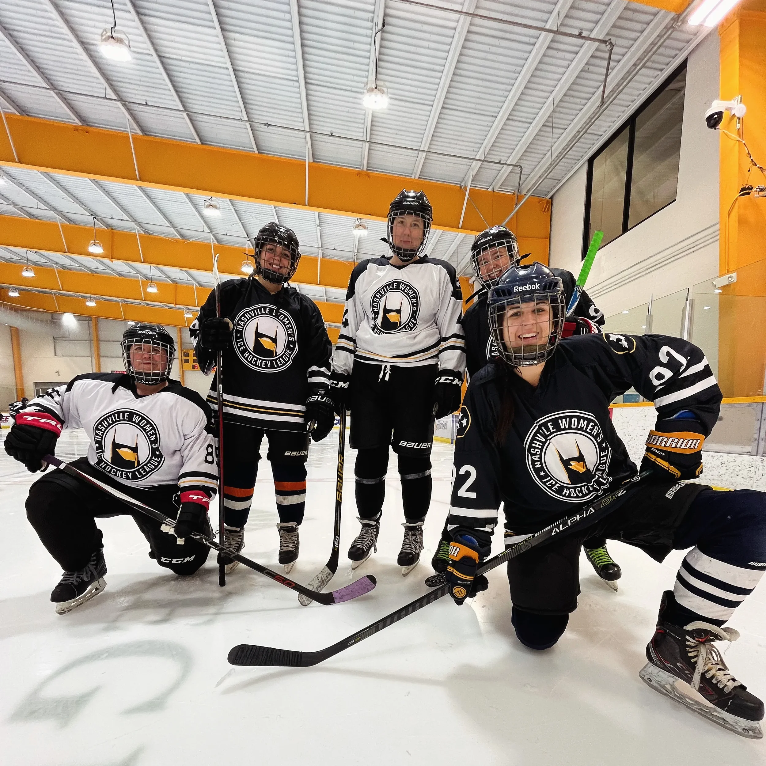 Five women in hockey gear posing on an ice rink, wearing jerseys with a logo that says 'Nashville Women's Ice Hockey League,' some kneeling and some standing, with hockey sticks and helmets.