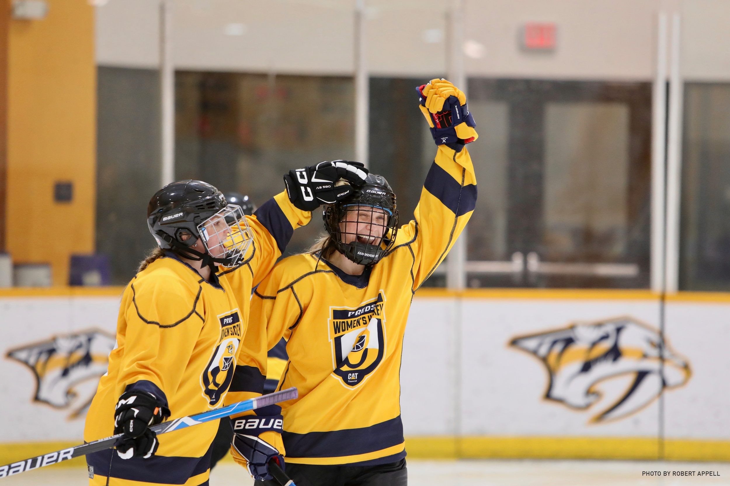 Two female ice hockey players in yellow jerseys celebrating on the ice rink, one raising her arm while the other is smiling and holding a hockey stick.