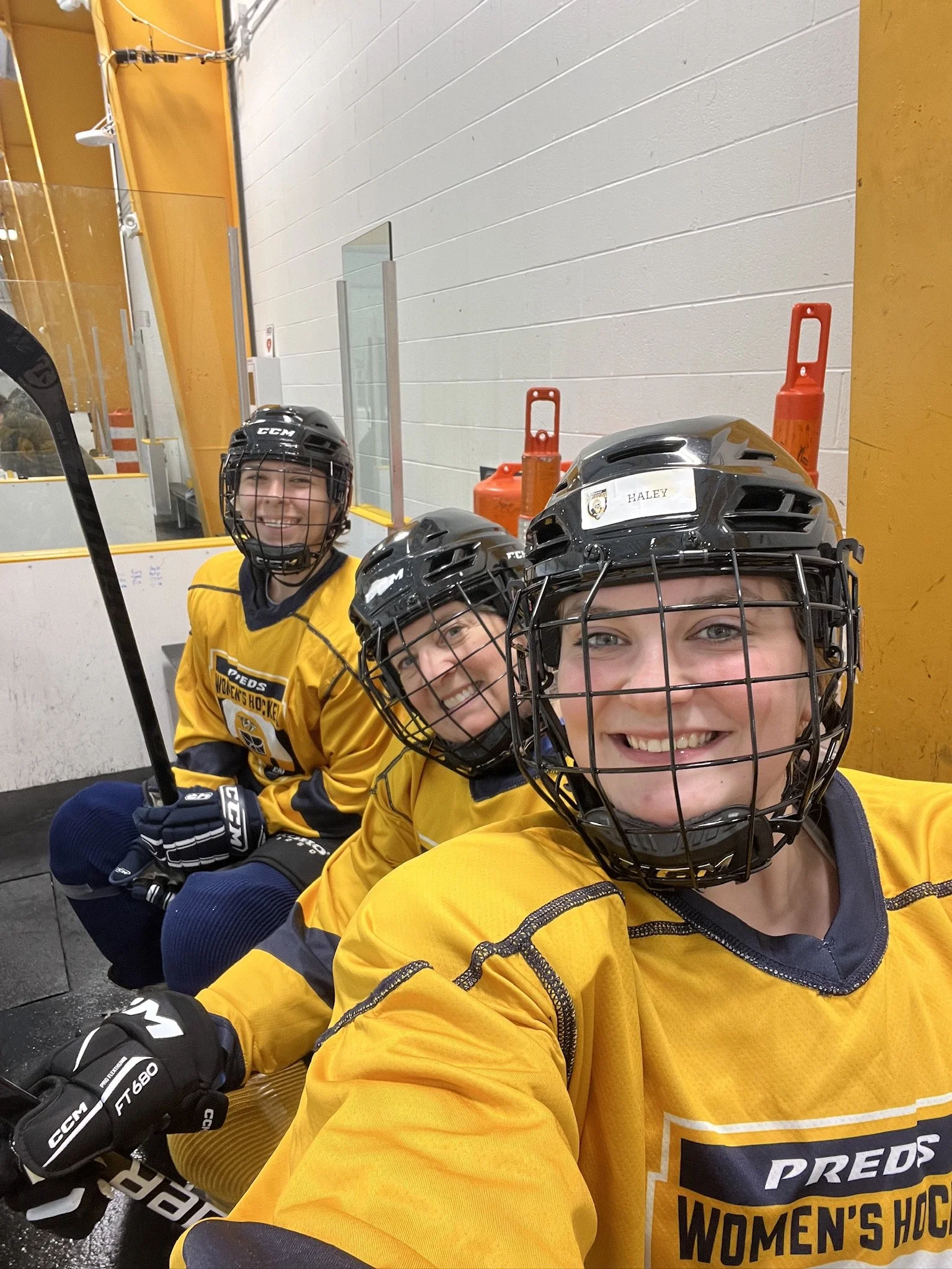 Three women in yellow hockey jerseys and black helmets sitting on a bench at an indoor hockey rink, smiling at the camera.