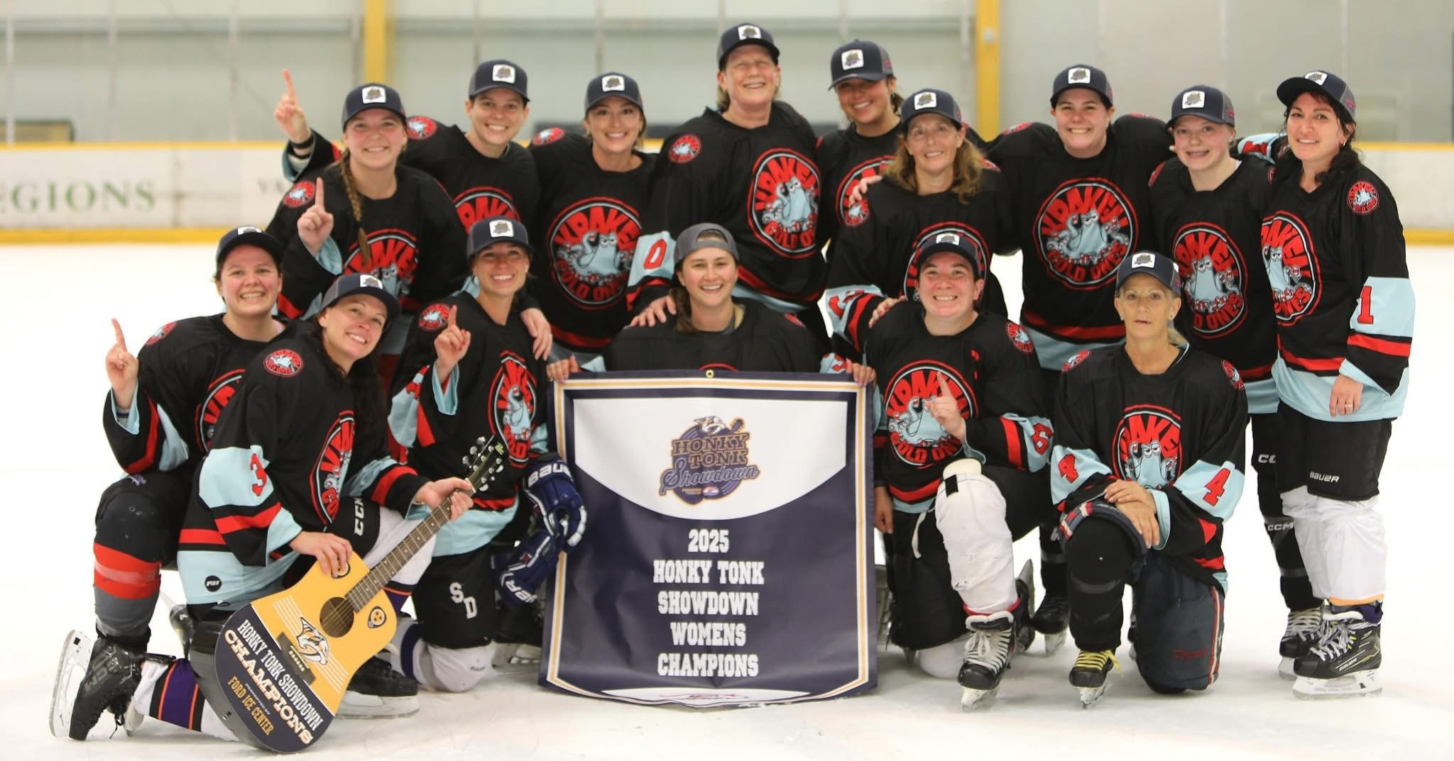 A women's ice hockey team celebrating their victory, posing on the ice rink holding a championship banner that reads '2023 Honky Tonk Showdown Women's Champions'. They are wearing black and light blue jerseys with a dragon mascot and caps, some playe