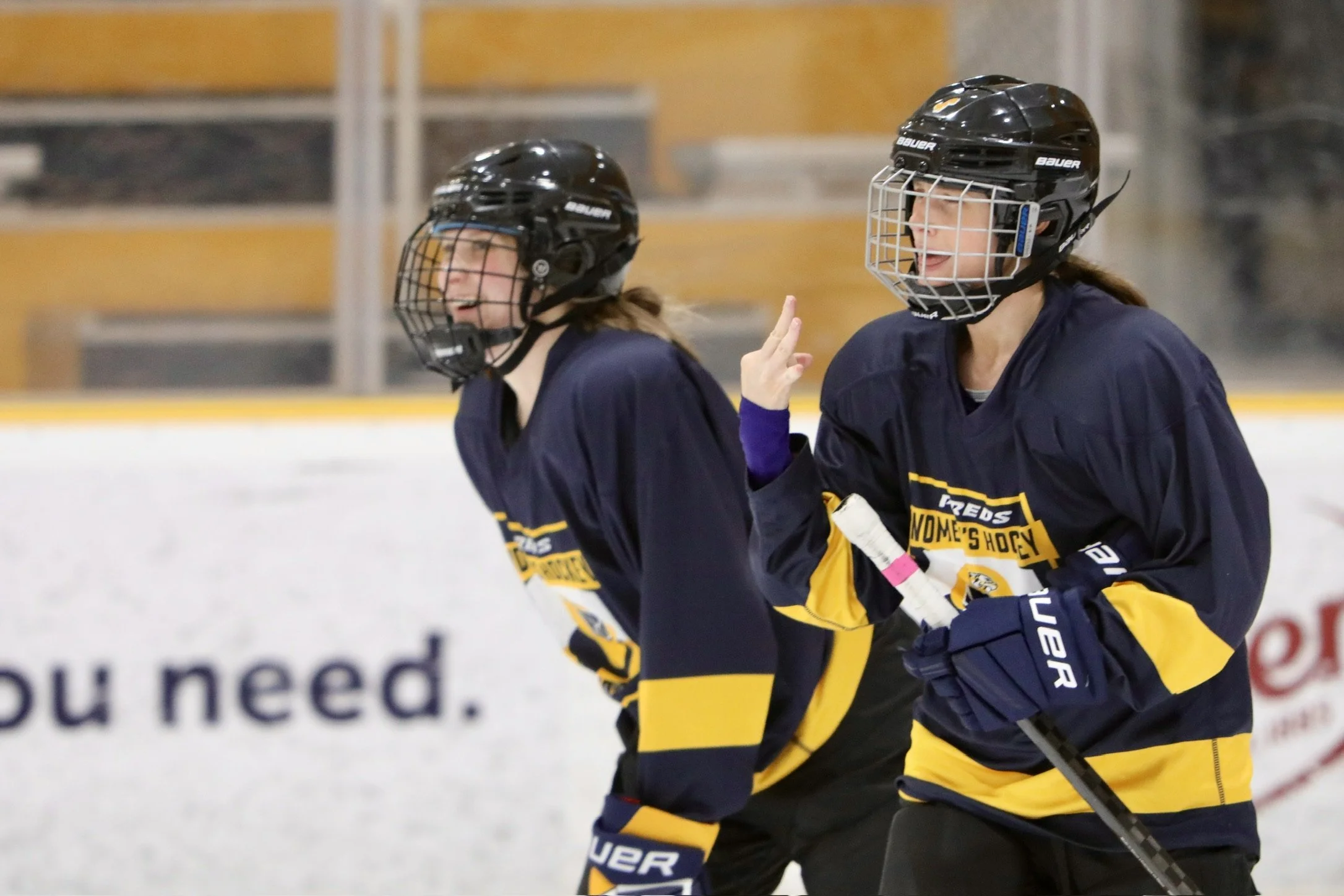 Two female ice hockey players in navy and yellow uniforms and black helmets, one of them is making a gesture with her hand, on an ice rink.