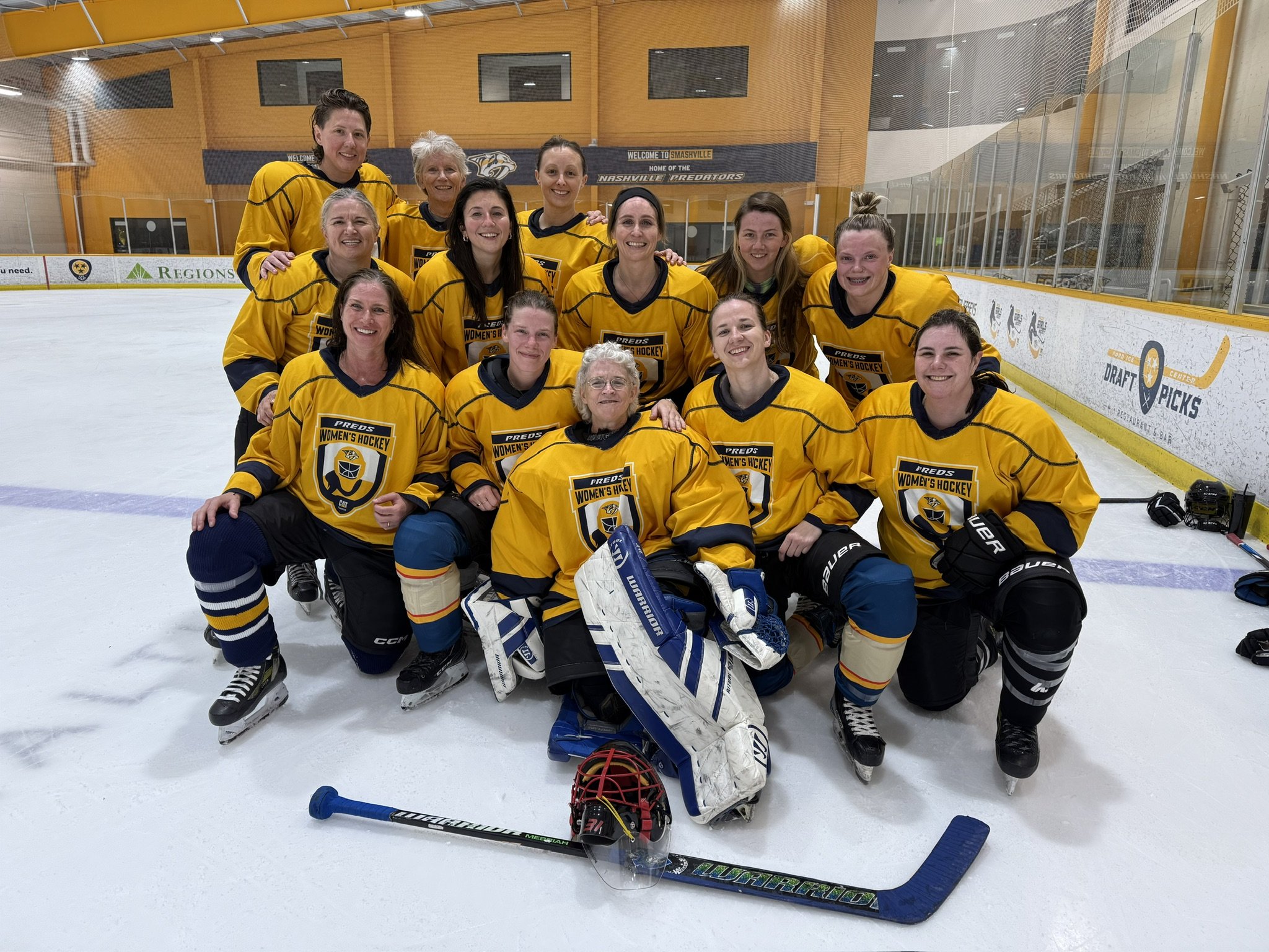 A women's hockey team in yellow jerseys posing on the ice rink with their coach, all smiling.