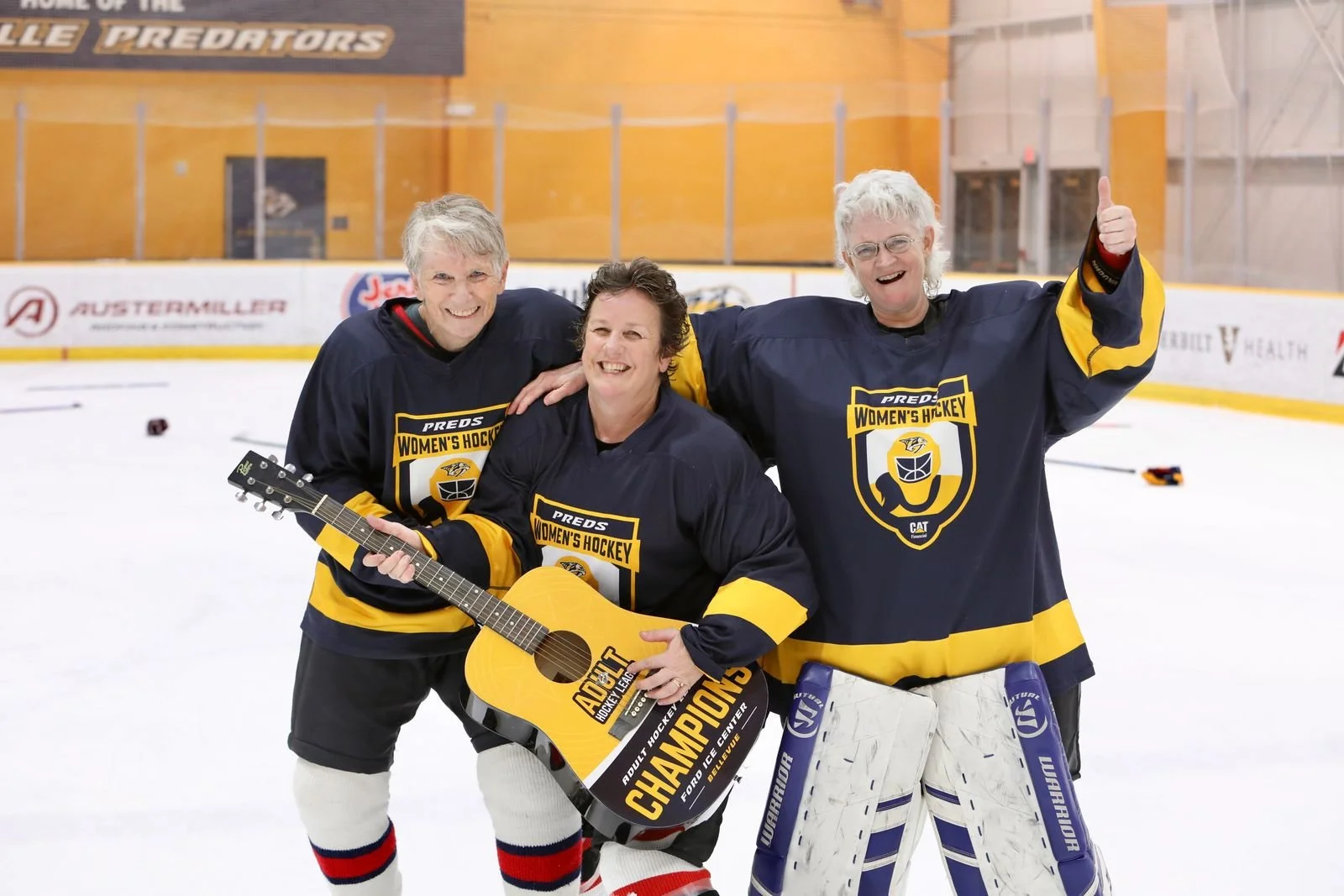 Three women on an ice hockey rink celebrating with one holding a guitar and a sign that says 'Champions.' They are wearing navy blue and yellow hockey jerseys that say 'Women's Hockey.' Two women are wearing white goalie pads, and all three are smili