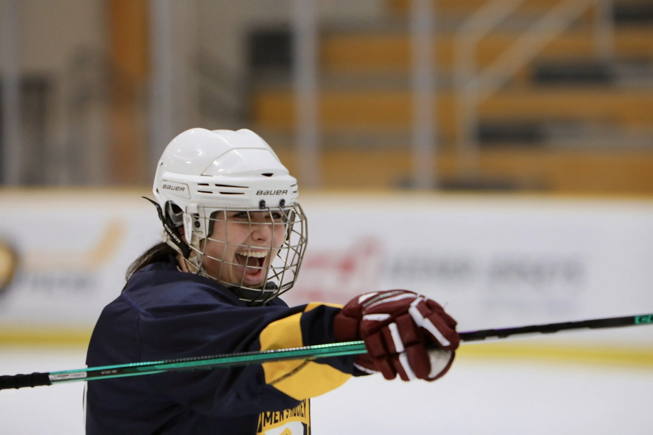 Female ice hockey player celebrating on the ice, wearing a white helmet with a cage, a navy and yellow jersey, and red gloves, holding a hockey stick.