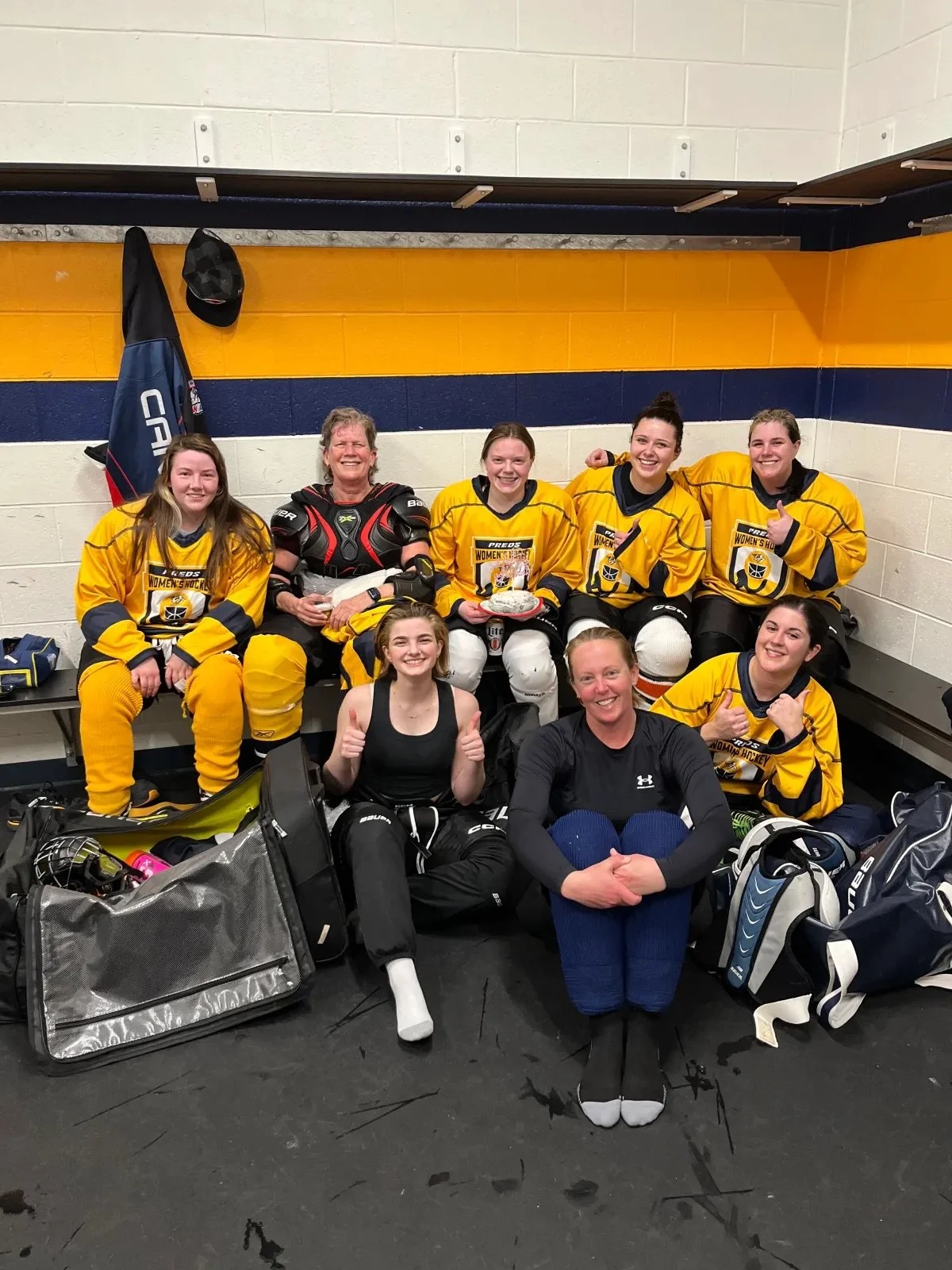 A women's hockey team sitting on a bench in a locker room, wearing yellow jerseys, with a coach or team staff member sitting in front.