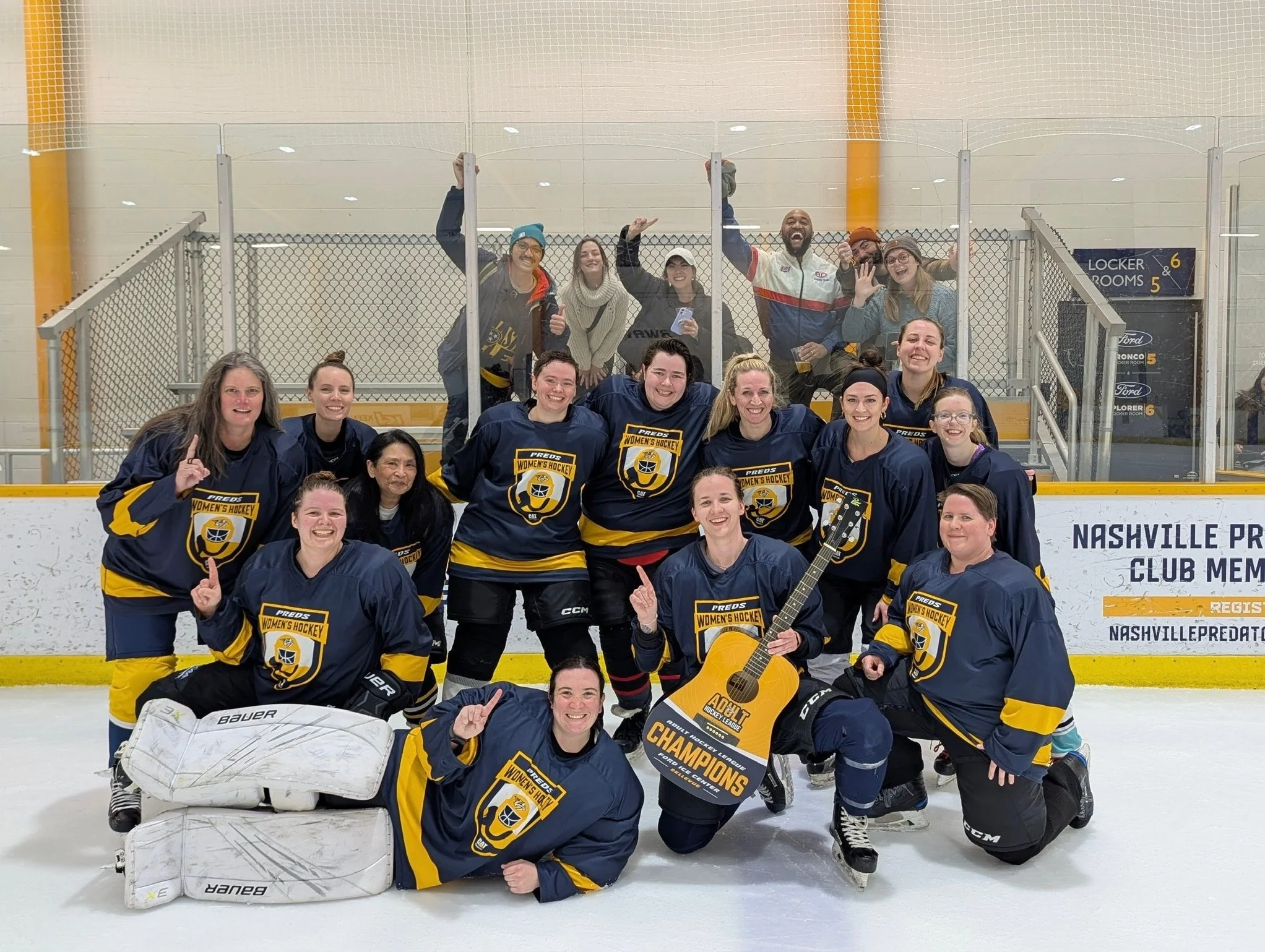 A group of women hockey players in navy and yellow uniforms celebrating on the ice rink, some holding a championship sign, with a few standing behind a clear protective barrier.