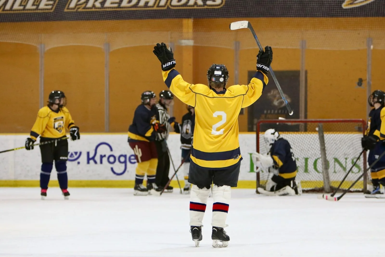 Ice hockey players on the ice rink, one player wearing a yellow jersey with the number 2 raising arms in celebration, others in dark jerseys, some near the goal, one goalie in white protective gear kneeling in front of the net.