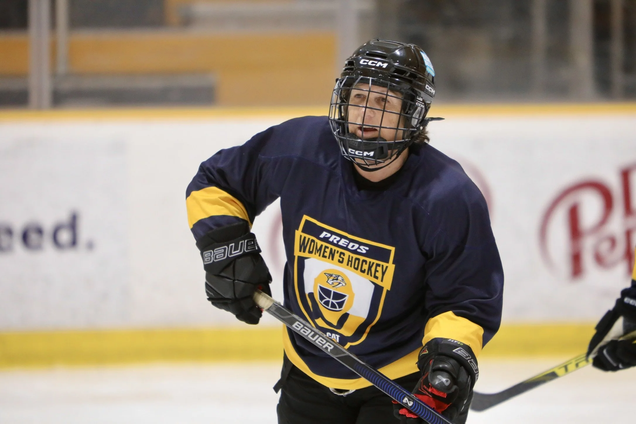 A female ice hockey player wearing a navy blue and yellow jersey with 'PREDS WOMEN'S HOCKEY' logo, black helmet, and gloves, holding a hockey stick on the ice during a game.