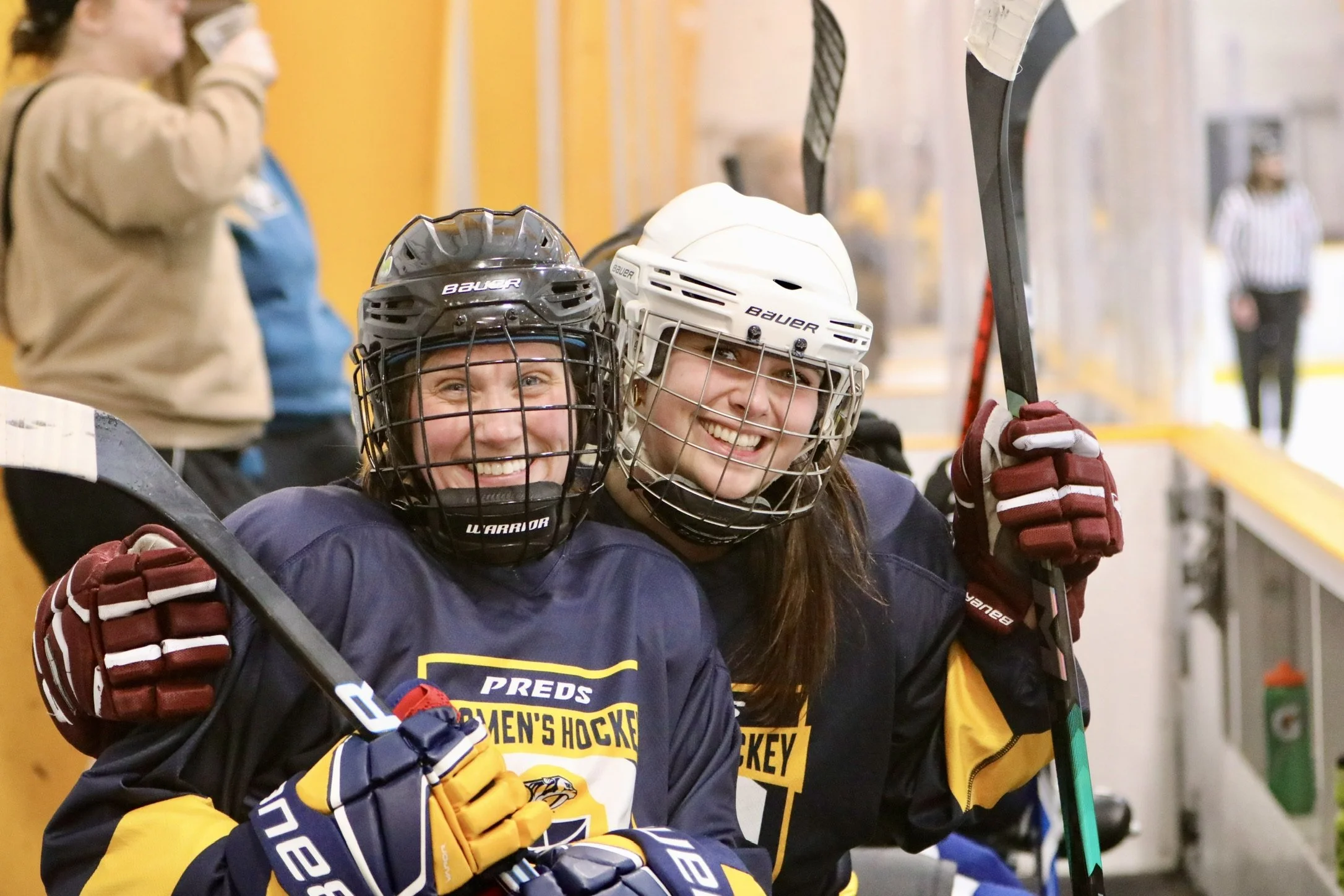Two female hockey players in full gear smiling and celebrating on the bench with their sticks