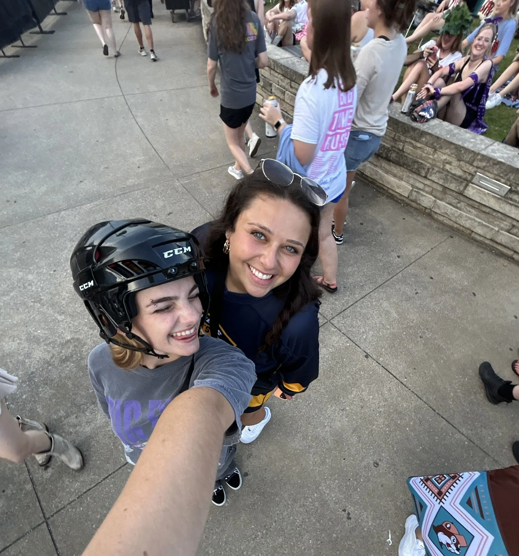 Two young women smiling for a selfie at an outdoor event, one wearing a hockey helmet and the other with sunglasses on her head, surrounded by a crowd of people sitting on a stone wall and standing around.