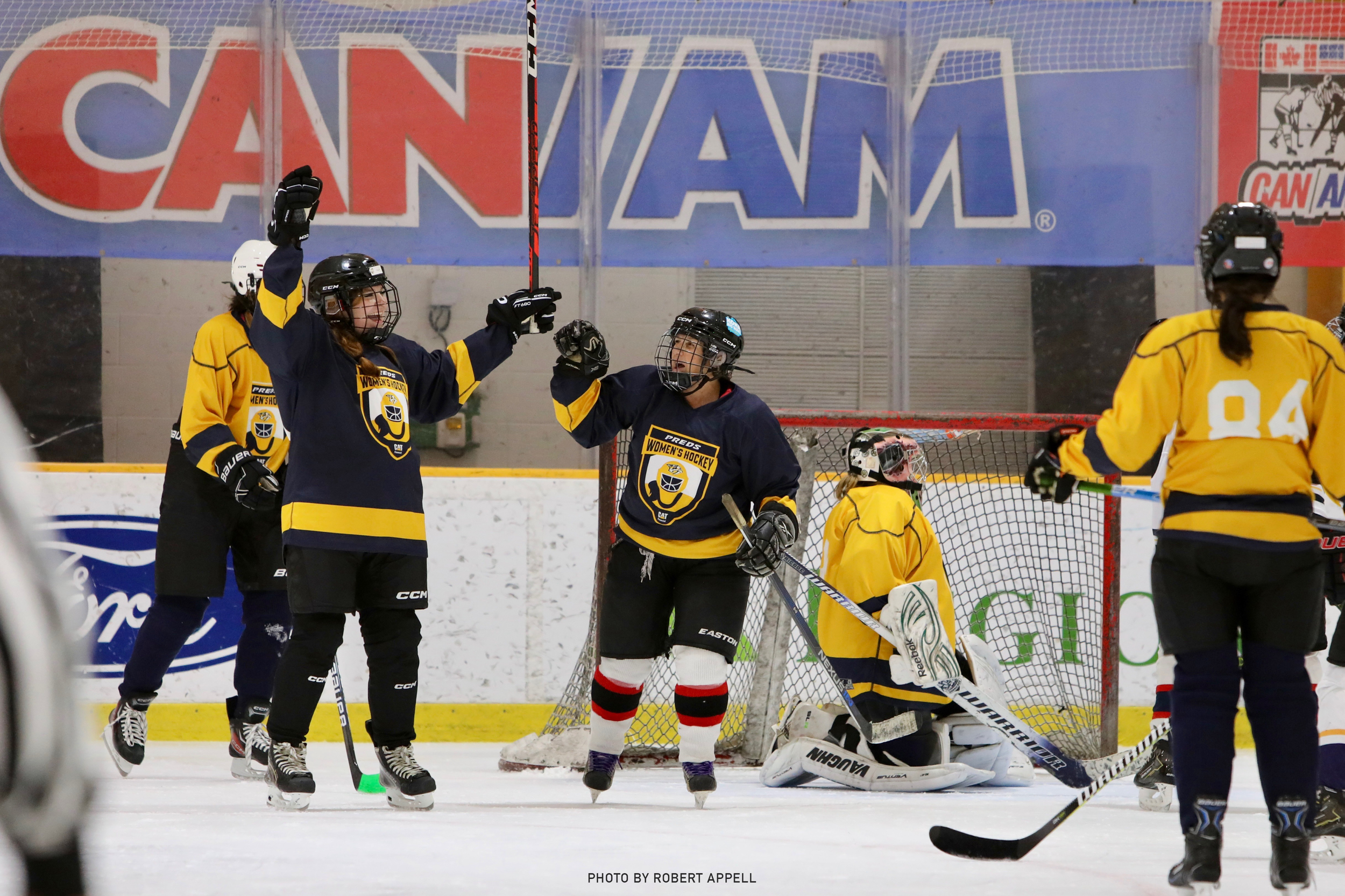 Women’s ice hockey players in yellow and navy uniforms celebrating on the ice rink, one with arms raised, with the goalie's net behind them.