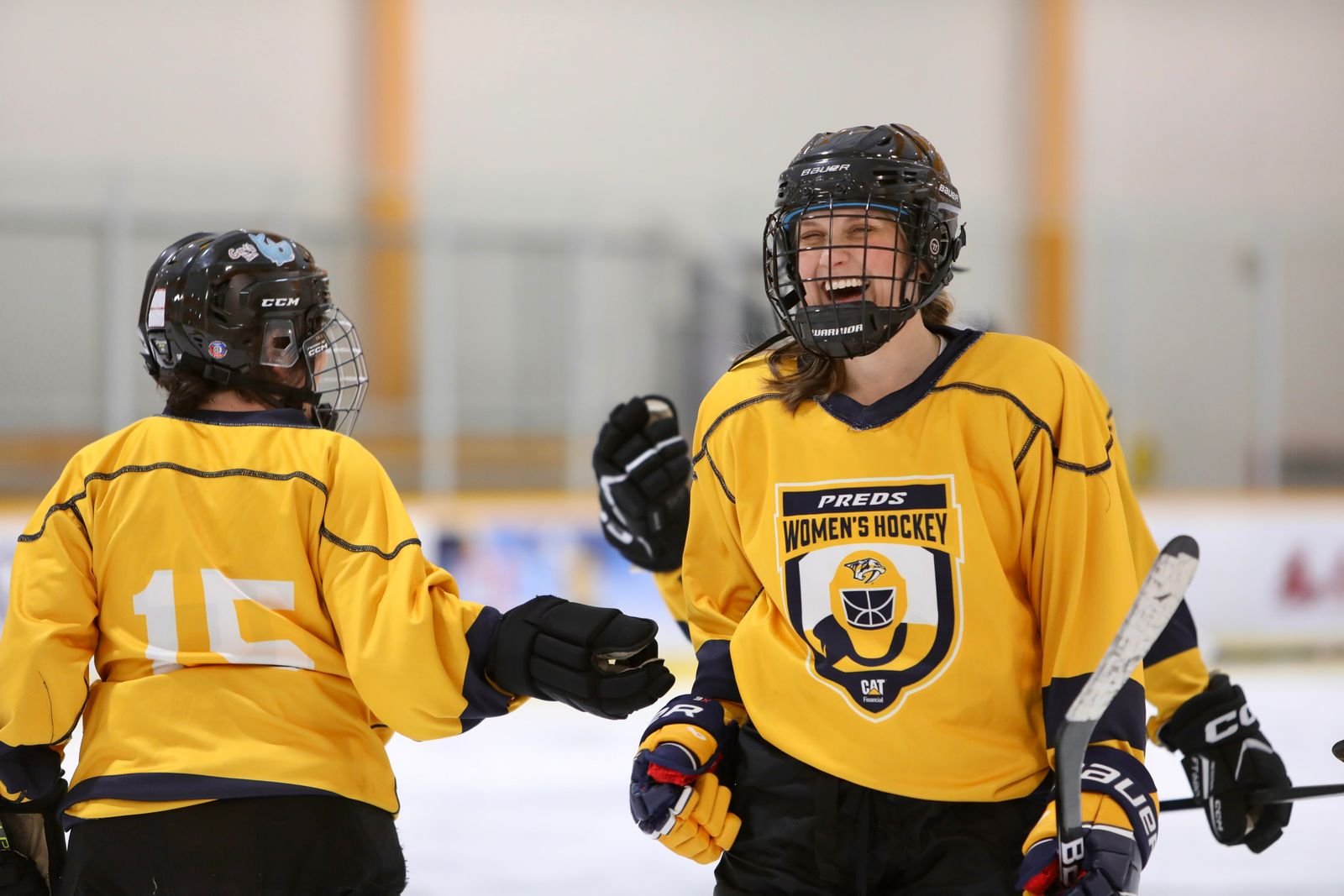 Two women hockey players in yellow jerseys celebrate on the ice rink, one smiling and the other giving a fist bump, with helmets and hockey sticks.