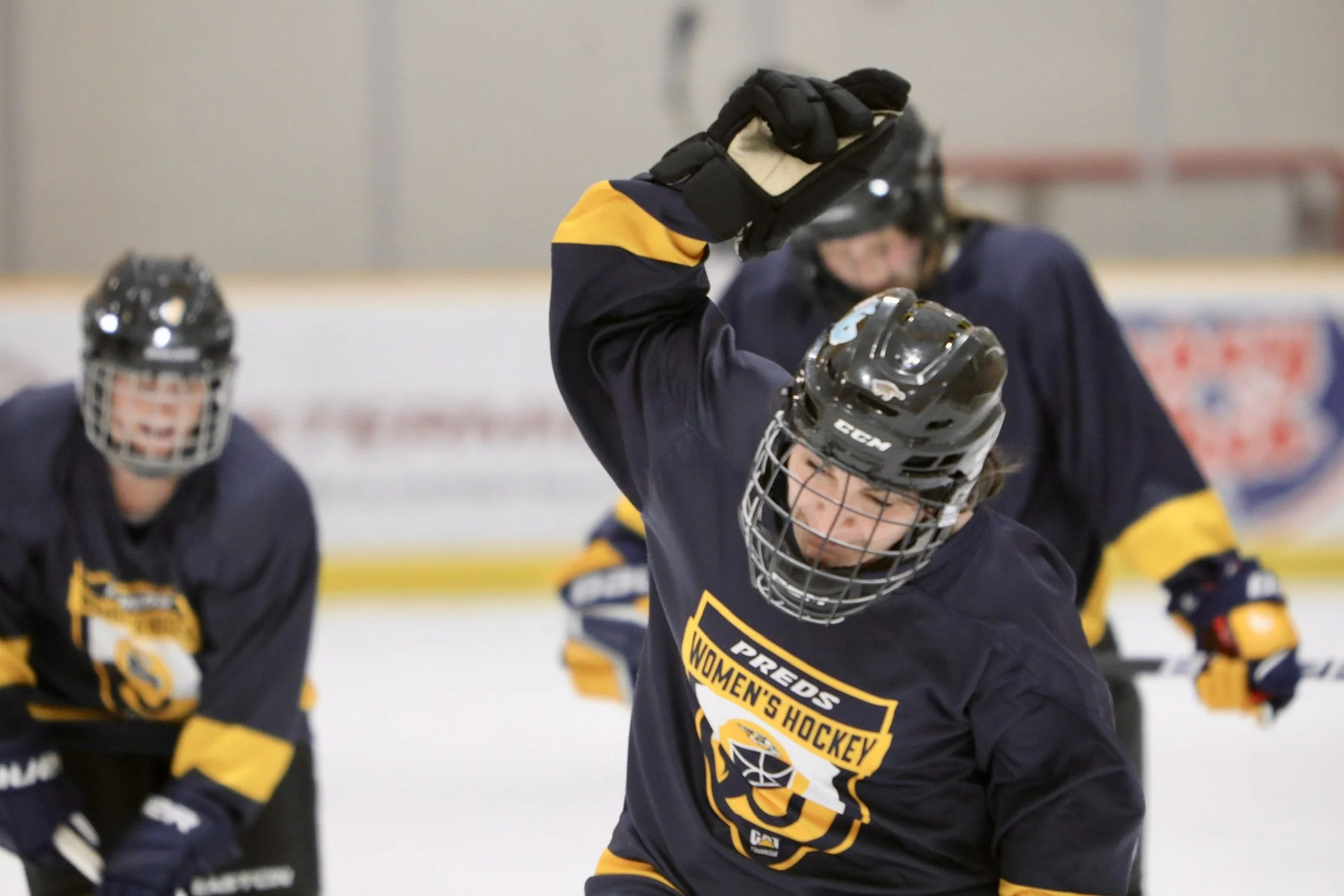 Female hockey players wearing black jerseys and helmets during a game at an ice rink, with one player raising her arm in celebration.