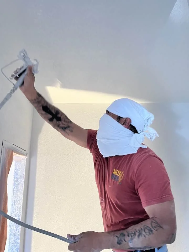 A person wearing a red shirt and a white cloth covering their head and face, using a sprayer to paint a ceiling.