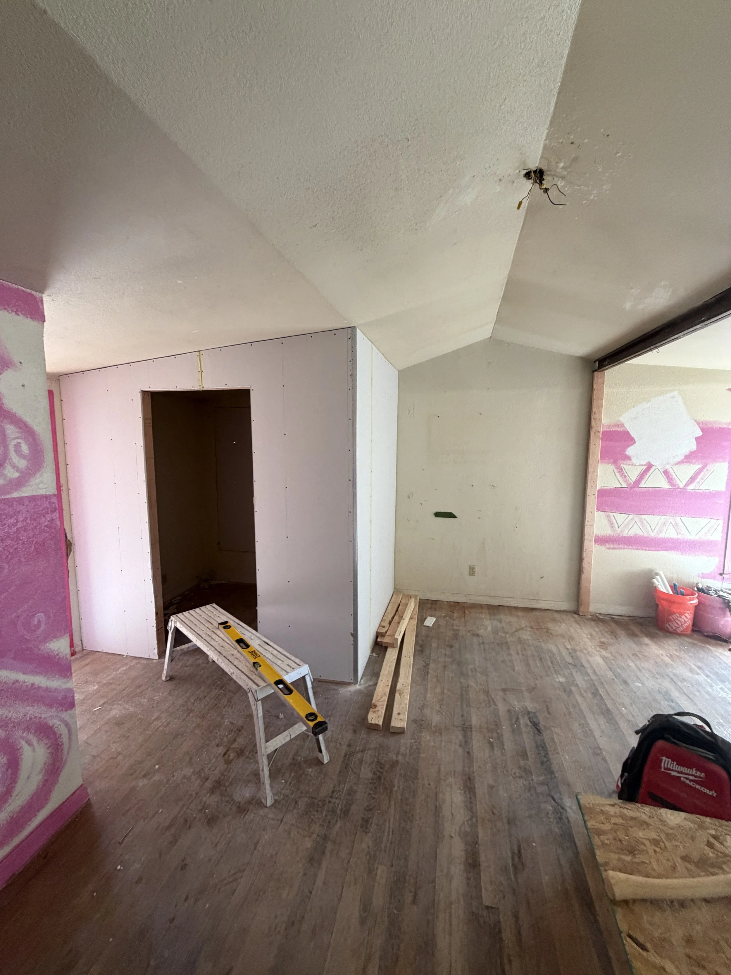 Interior of a room under renovation with drywall partially installed, wooden planks and construction tools on the floor, and a ladder.