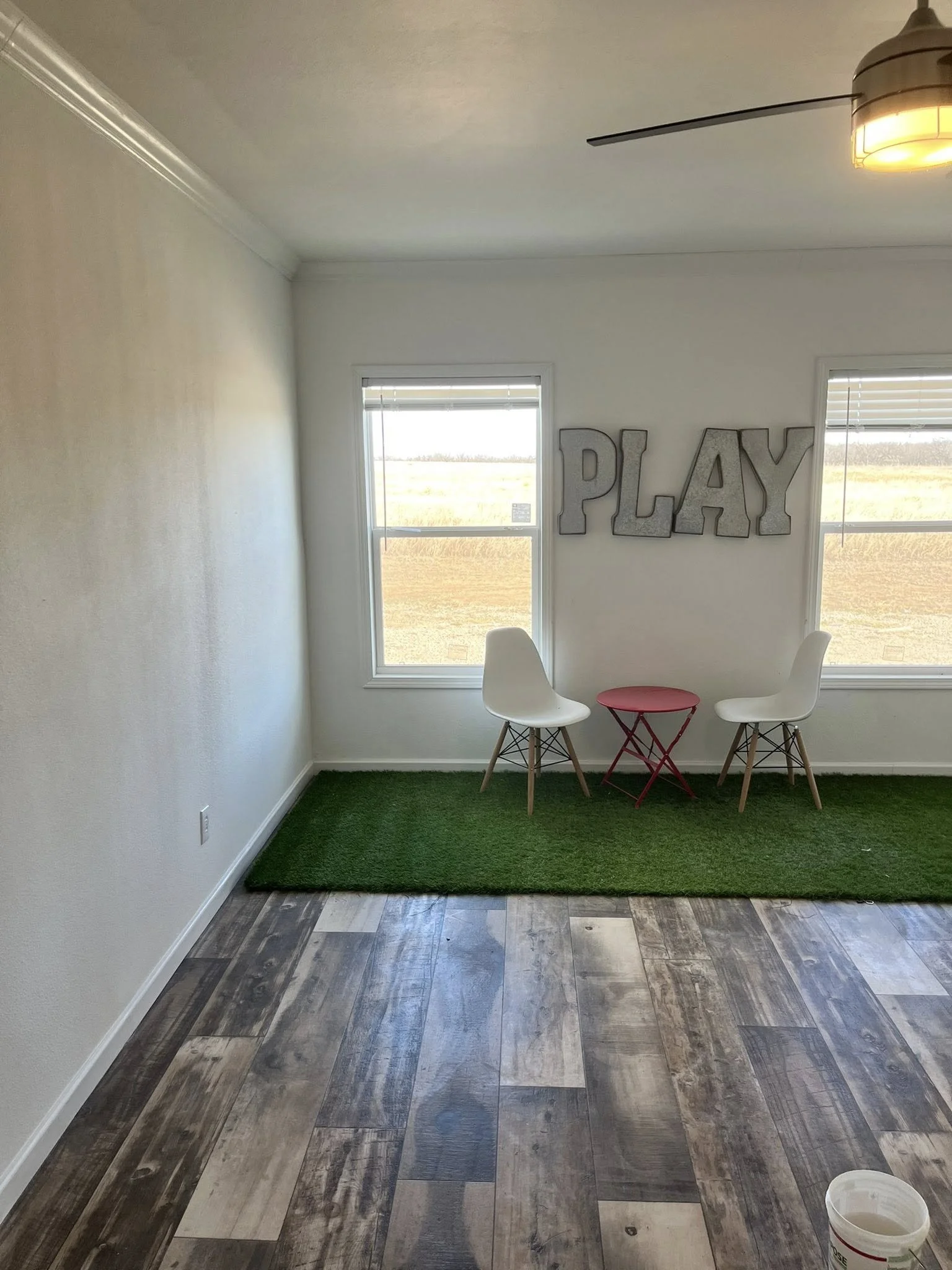 Interior of a room with two windows, a white wall with large metal letters spelling 'PLAY' hanging on it, two white chairs, a small red table, a green rug, wooden floor, and ceiling fan with light.
