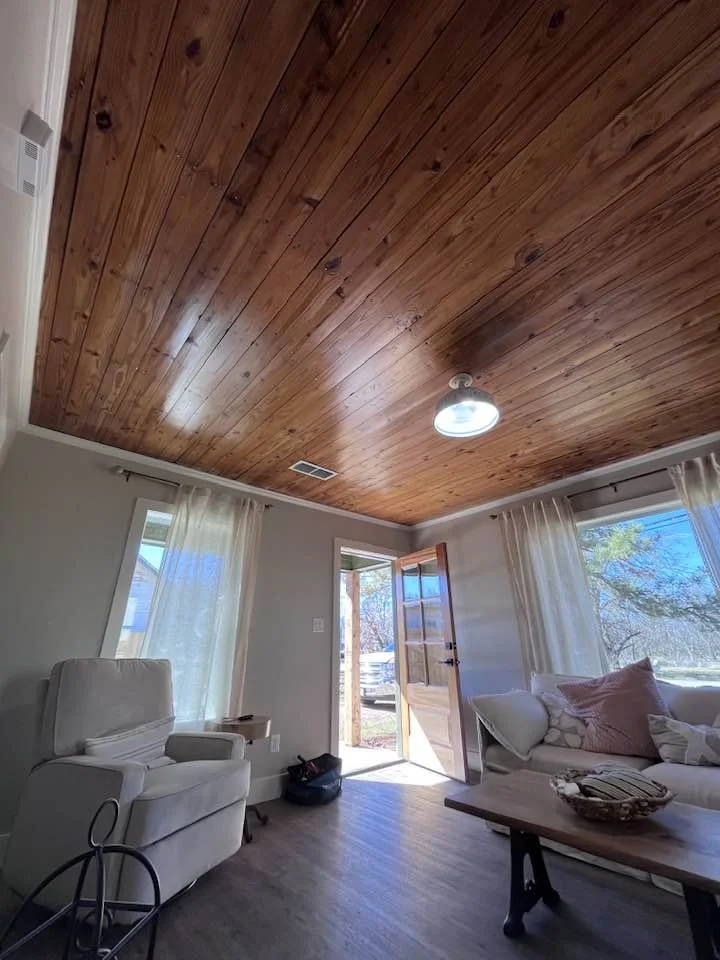 Living room with wooden ceiling, light-colored walls, two large windows with curtains, an open door to the outside, a white armchair, a sofa with cushions, a coffee table, and natural sunlight.
