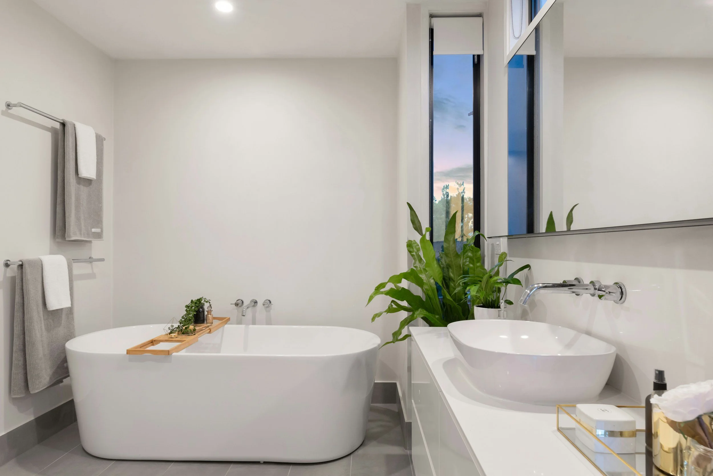 Modern white bathroom with a bathtub, green plants, a large mirror, and a window showing a sunset sky.