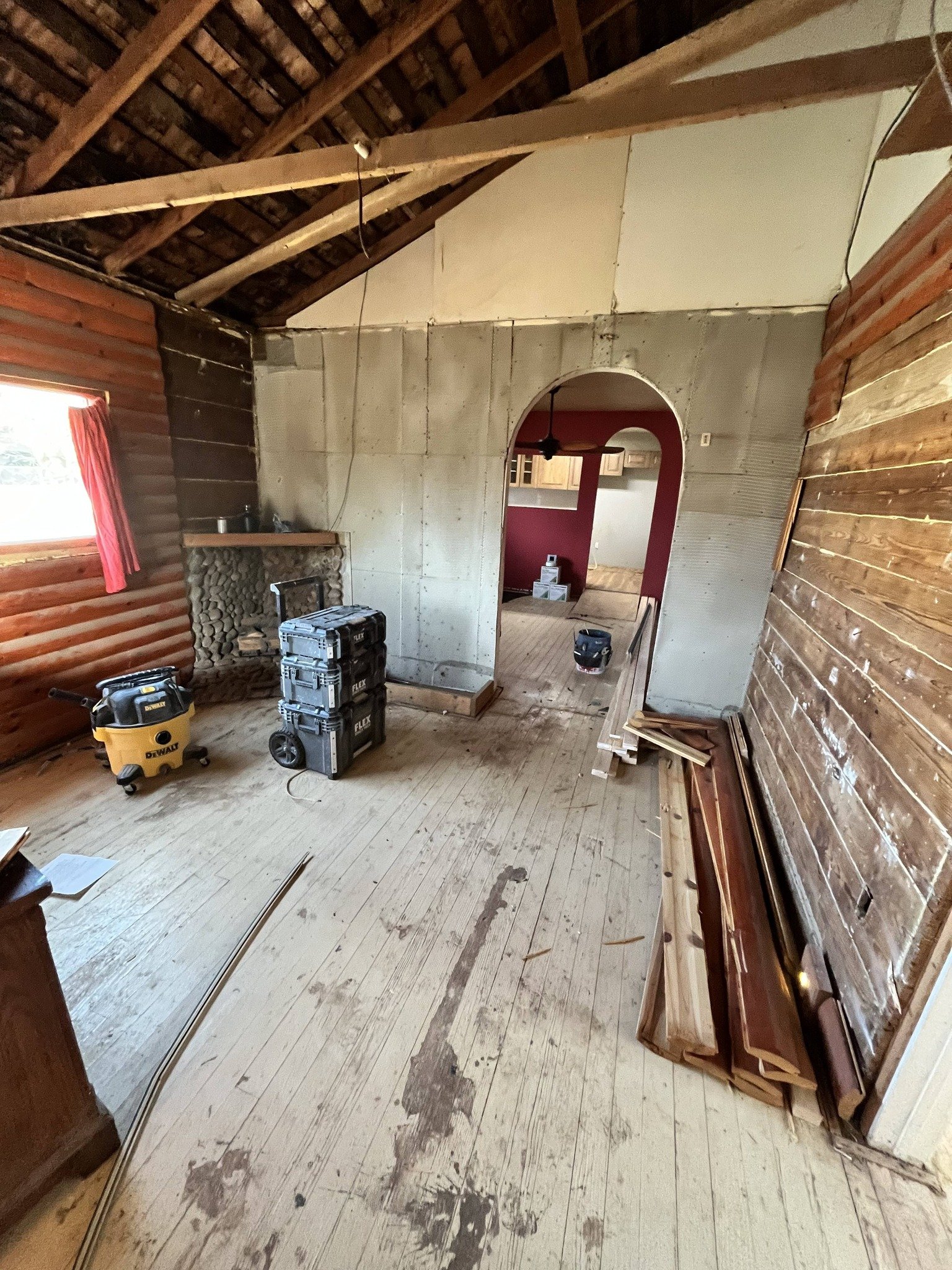 Interior of a room under renovation with unfinished walls, wooden flooring, construction tools, wood planks, and an arched doorway leading to another room with red walls.