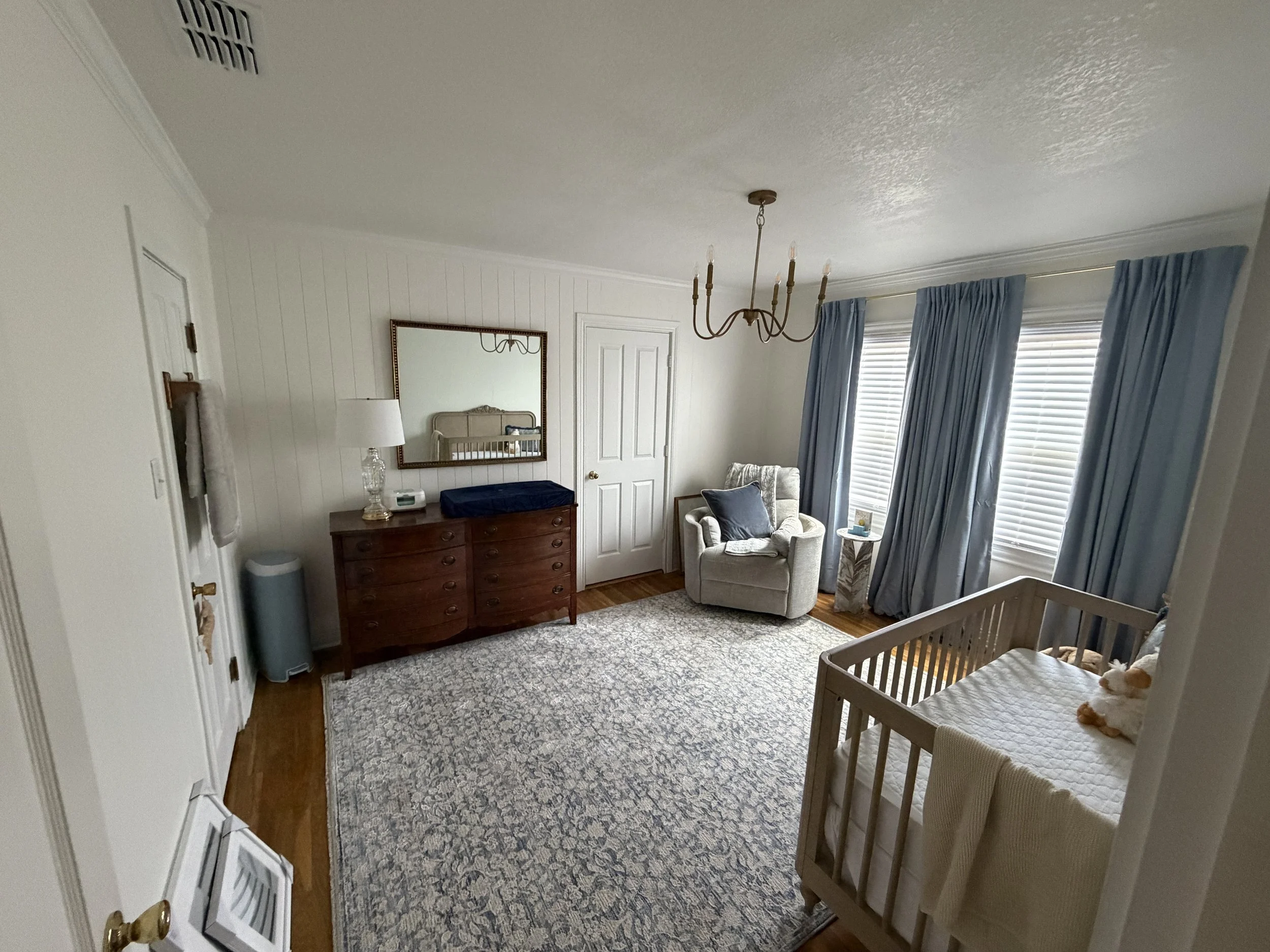 A cozy living room with light-colored walls, a white door, large windows with blue curtains, a chandelier, a mirror, a beige armchair with a throw pillow, a wooden dresser, a table lamp, a crib with a stuffed animal, and a patterned area rug.