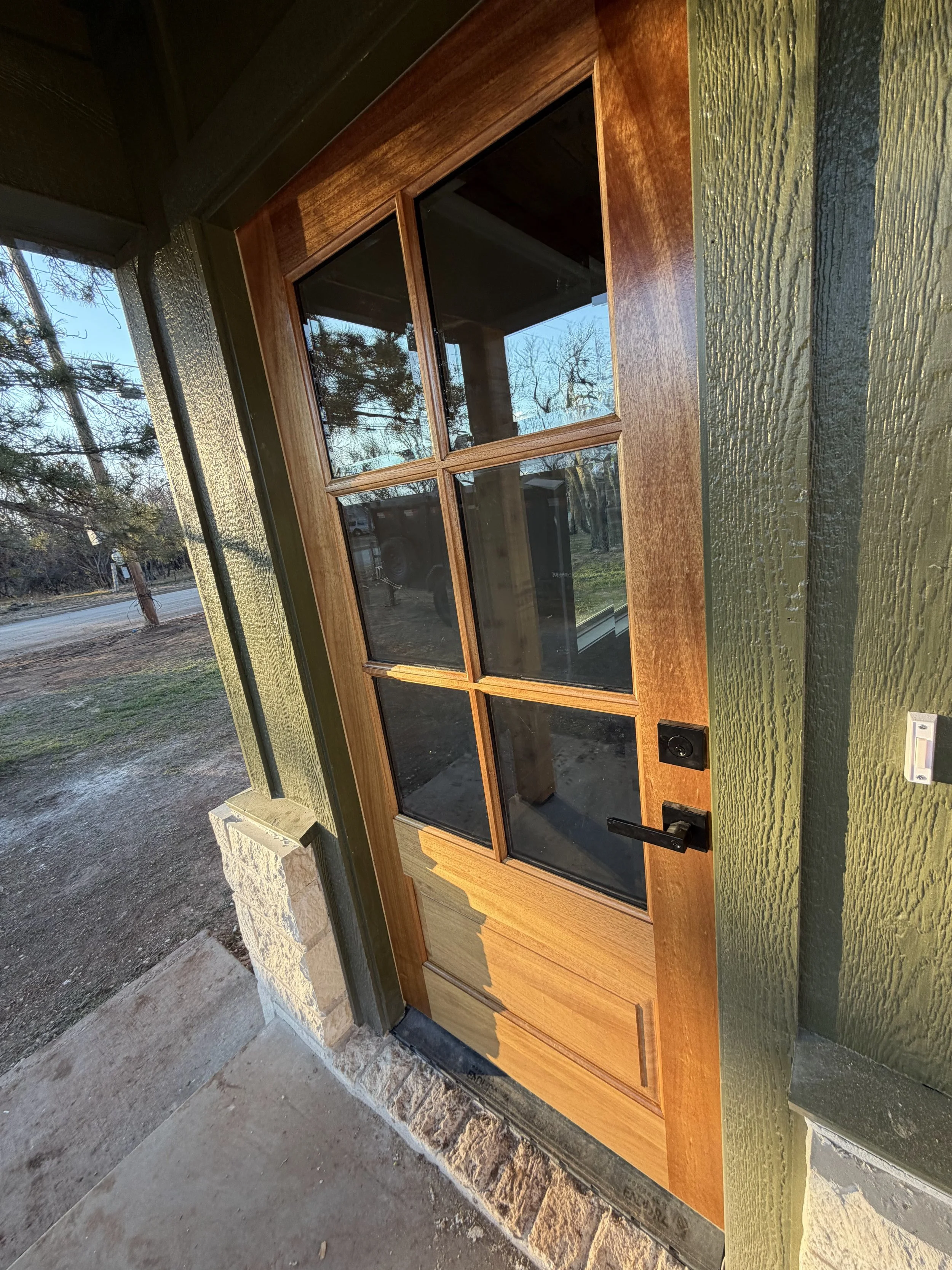 A front door made of wood with glass panes, set in a green-painted exterior wall. The door has a black handle and a deadbolt lock, with a stone step at the entrance.