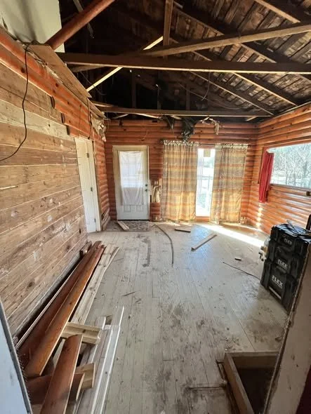 Interior of a rustic wooden cabin room under construction with exposed wooden beams, a door, a window with checkered curtains, various construction materials on the floor, and a black tool case.